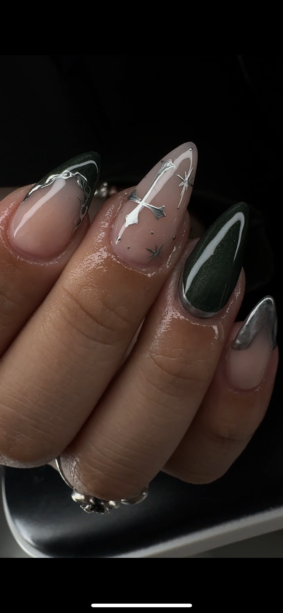 Close-up of a hand with decorative nail art. The nails are long and pointed, featuring designs including metallic silver accents, a white star and moon motif on a nude base, and dark green with white outlining.