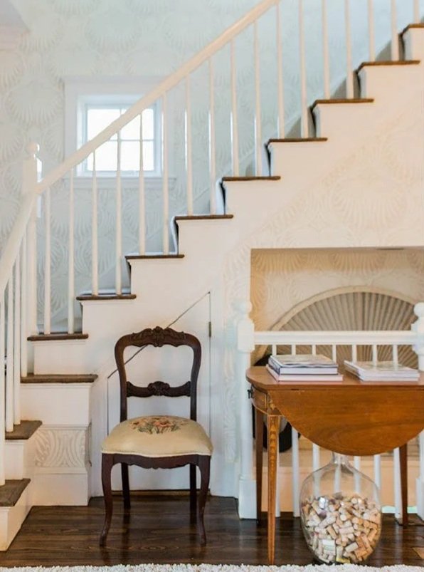 Indoor scene of a staircase with white railings and dark treads, a vintage wooden chair with floral upholstery, a small table with books, and a glass jar filled with wine corks, against patterned wallpaper.