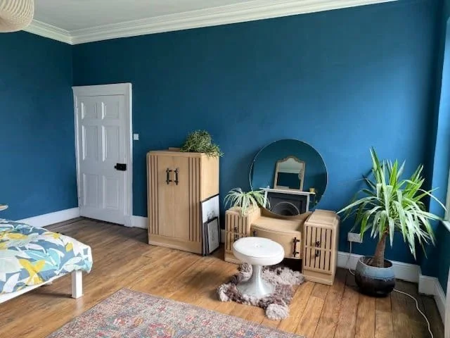 Living room with blue wall, white door, a wooden cabinet with green plant, round white table with fur rug, large potted plant, and bed partially visible on the left.