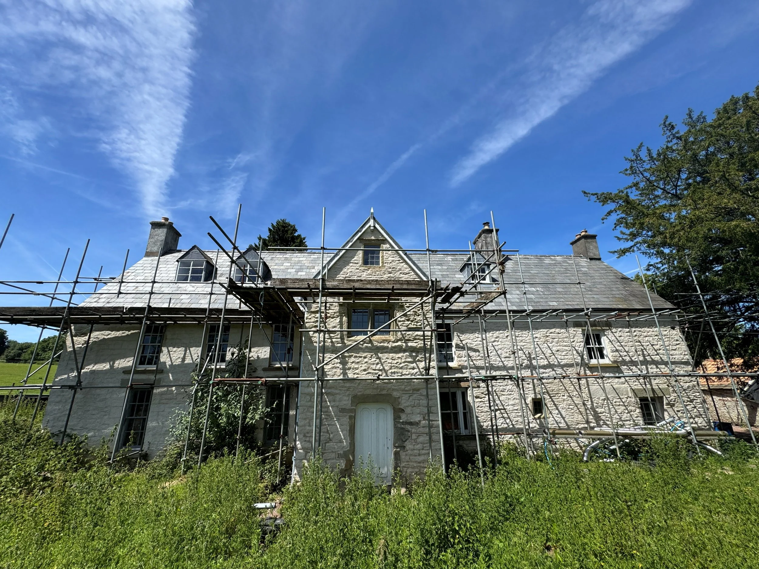 A stone house with a steep slate roof under construction, surrounded by metal scaffolding, set against a bright blue sky with wispy clouds and a large tree to the right.