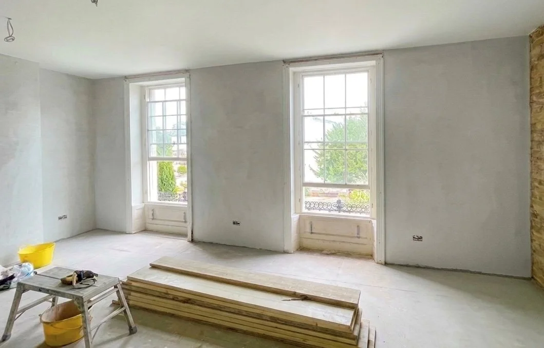 Inside a room under construction with two large windows, a small table with tools, and stacked wooden planks.