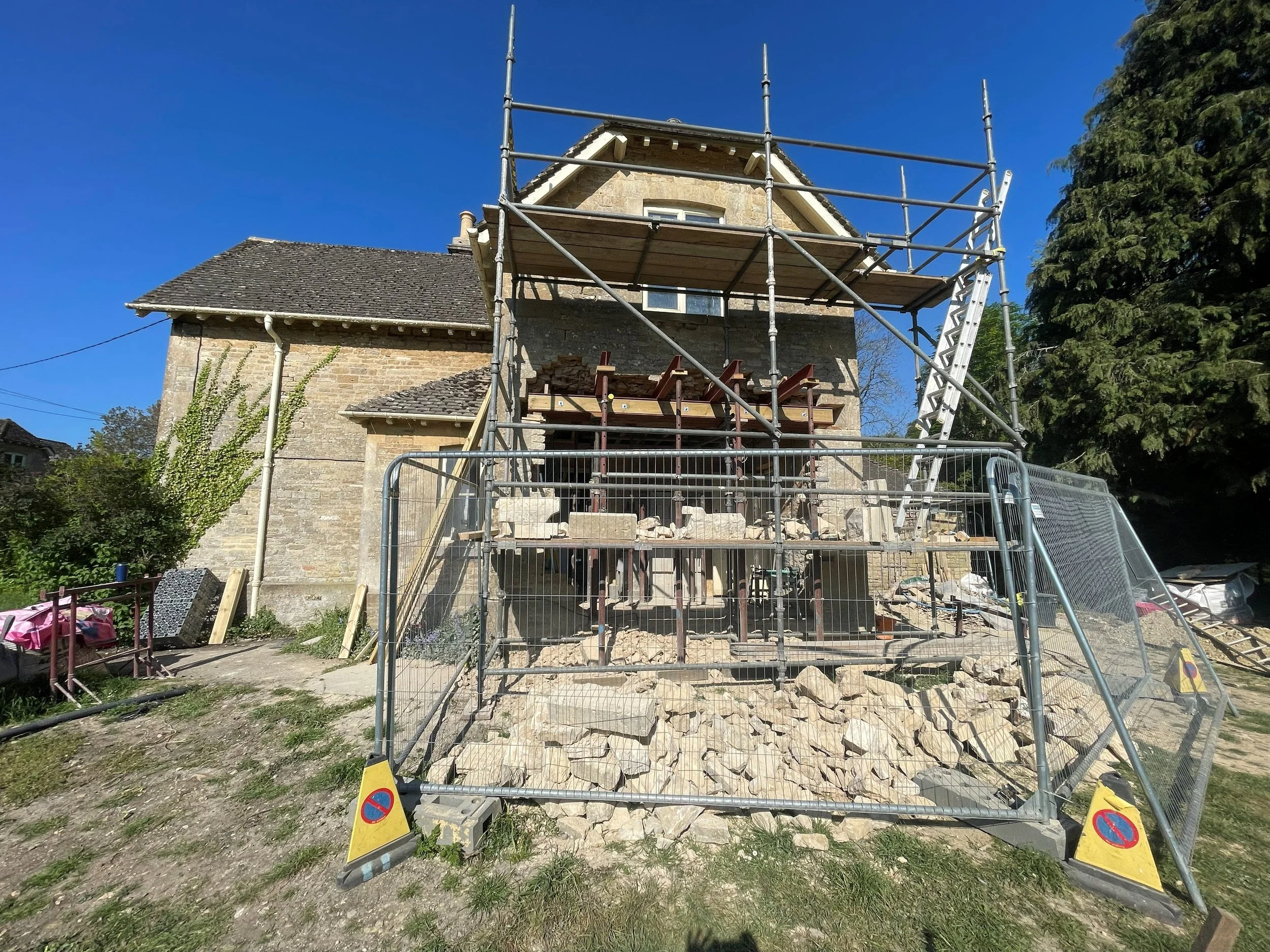 A house under renovation with scaffolding and a safety fence around a section of the building, rubble and construction materials at the base of the structure, and a clear blue sky overhead.