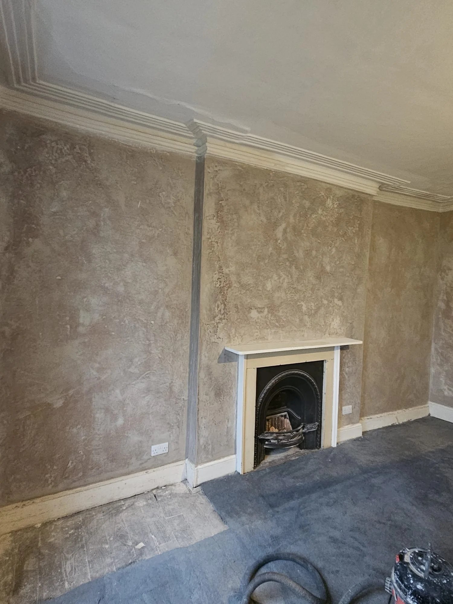 Living room with a decorative fireplace, partially finished wall with wallpaper or textured paint, and a vacuum cleaner on the carpet.