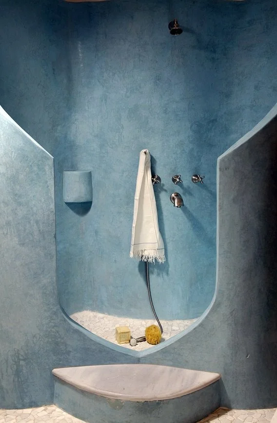 A shower with blue walls and a built-in ledge holding decorative items, a white towel hanging on hooks, and a rounded marble seat.