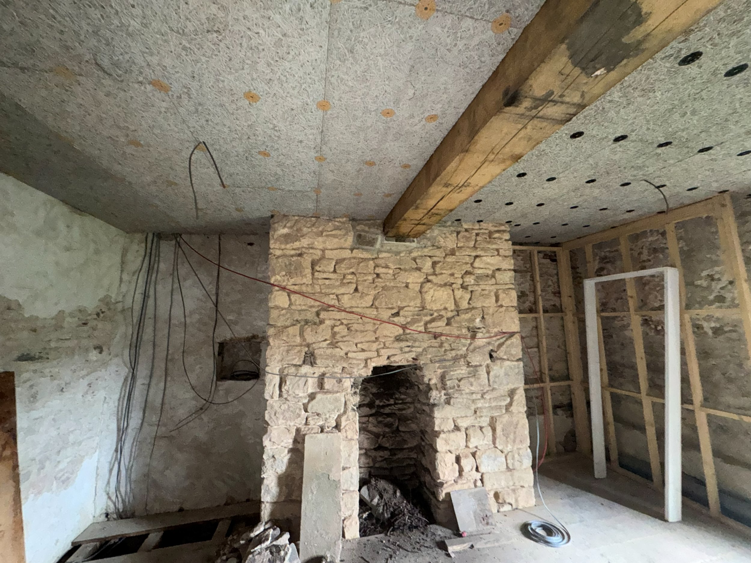 Construction site with stone wall and wooden beams, wires hanging from the ceiling, and construction materials in an unfinished room.