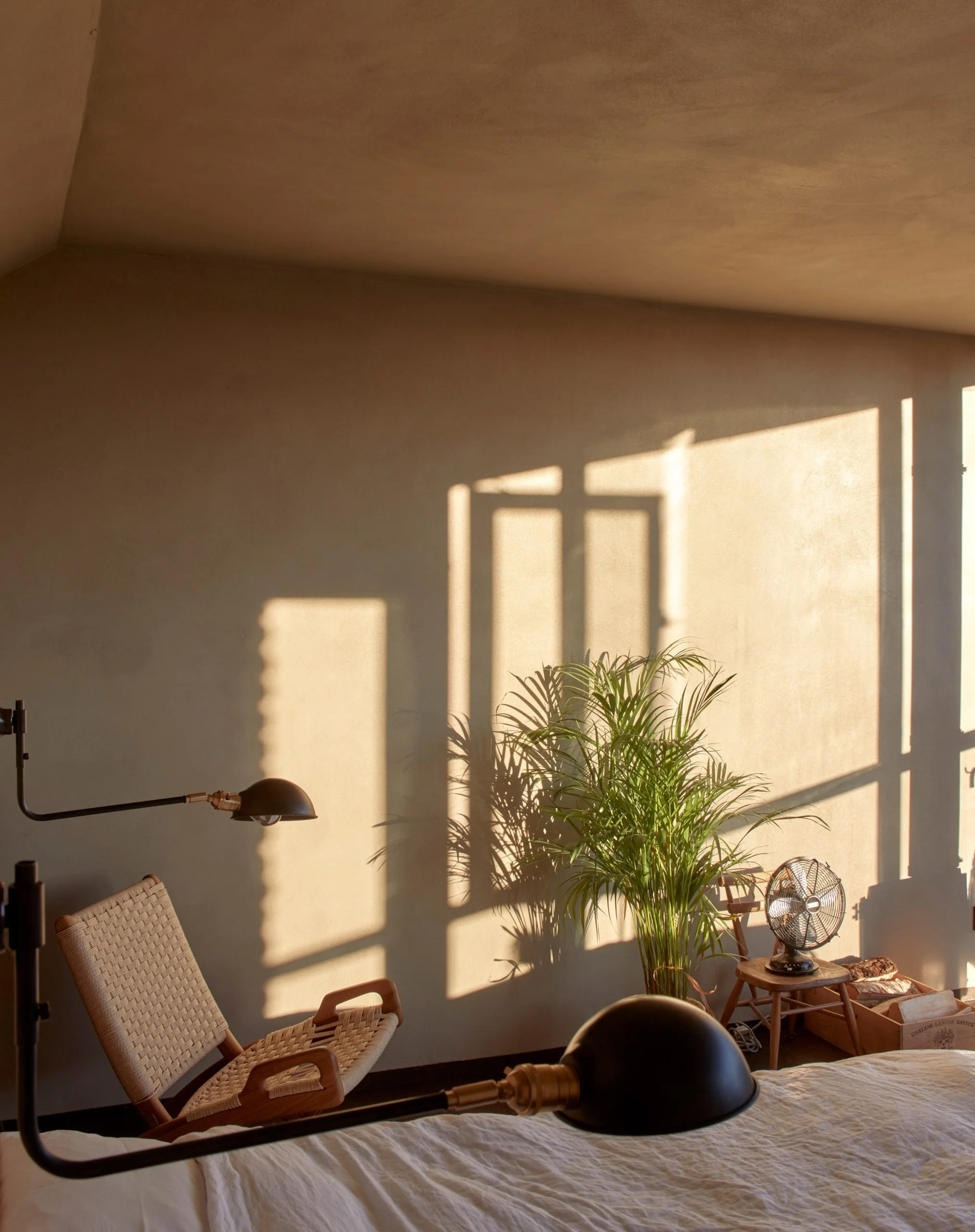 A cozy bedroom scene with sunlight casting shadows through a window onto a wall, a potted plant, a modern black bedside lamp on a wooden table, a woven chair, and part of a bed with a white coverlet.