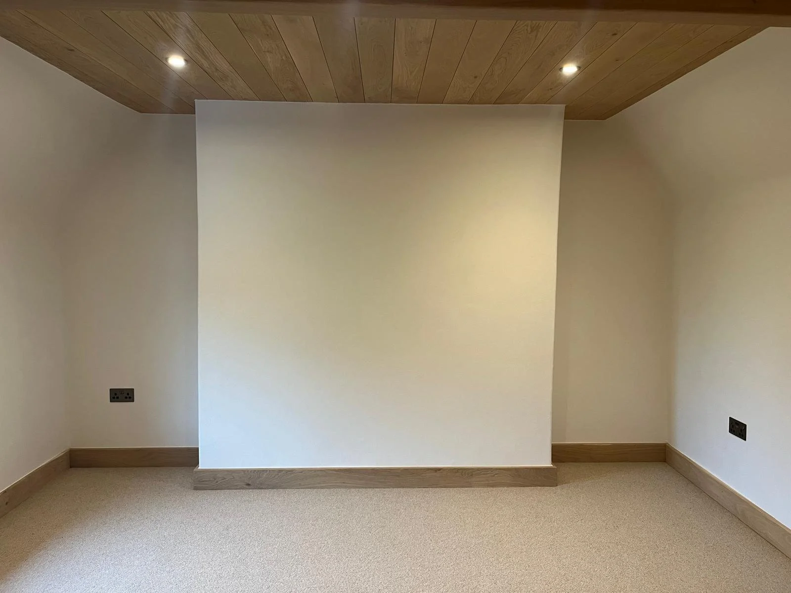 Empty corner of a room with white walls, a ceiling with wooden paneling, and beige carpeted floor. Two electrical outlets are visible on the walls.