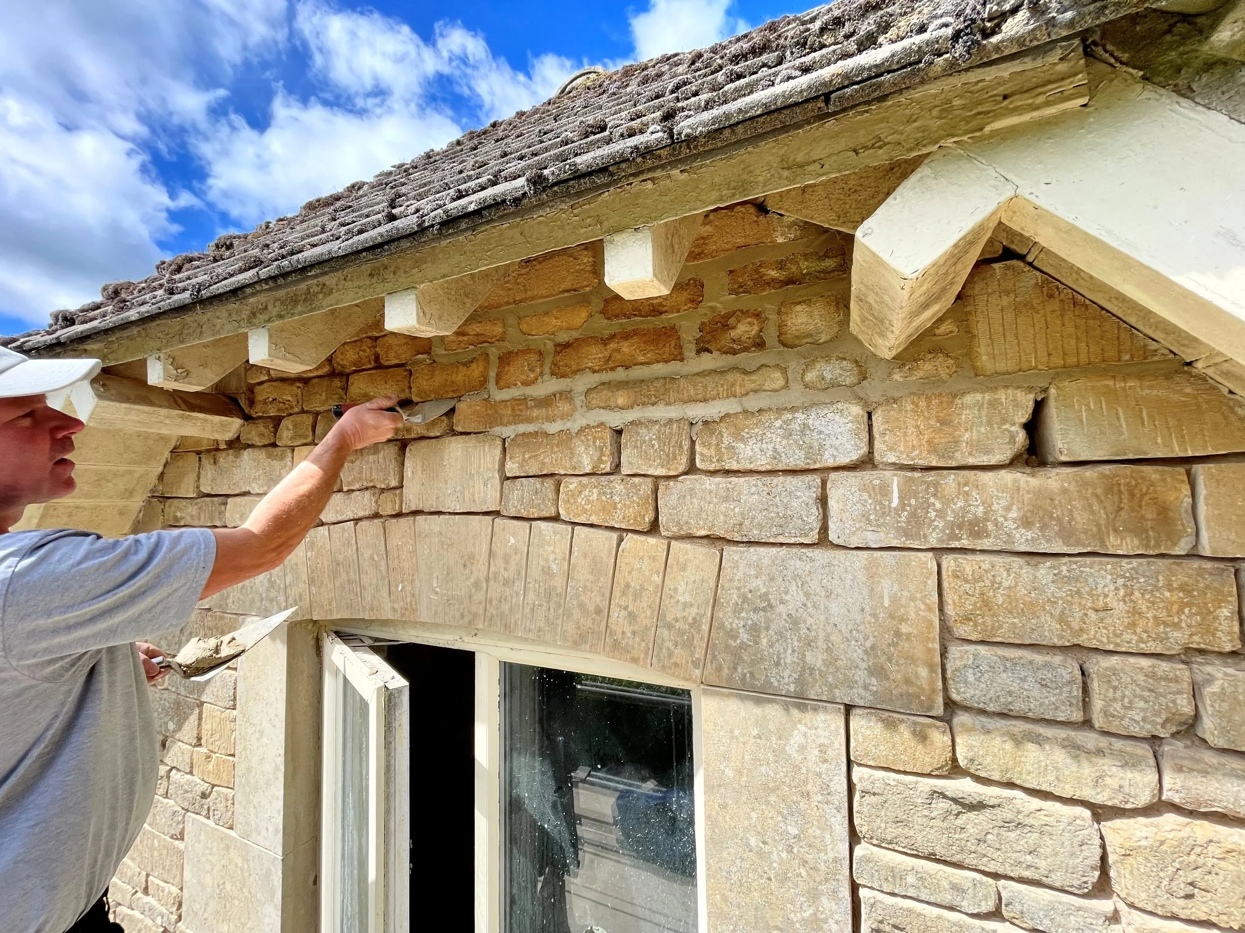 A person wearing a gray t-shirt and a white baseball cap is cleaning the soffit of a stone house with a tool, standing outside beneath the eaves under a partly cloudy sky.
