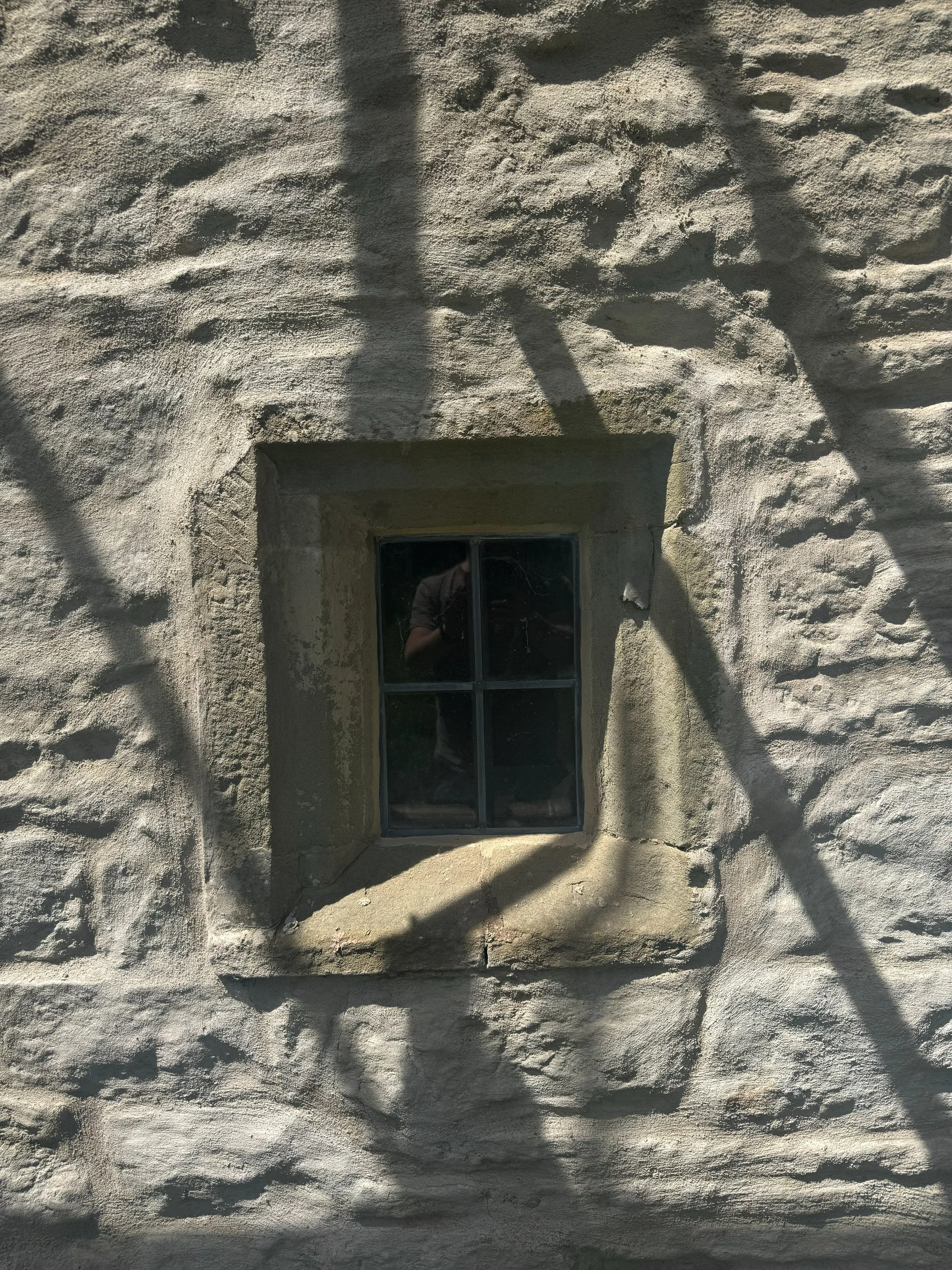 A textured beige stucco wall with a small rectangular window and a person's reflection visible in the glass, surrounded by shadowy tree branches.