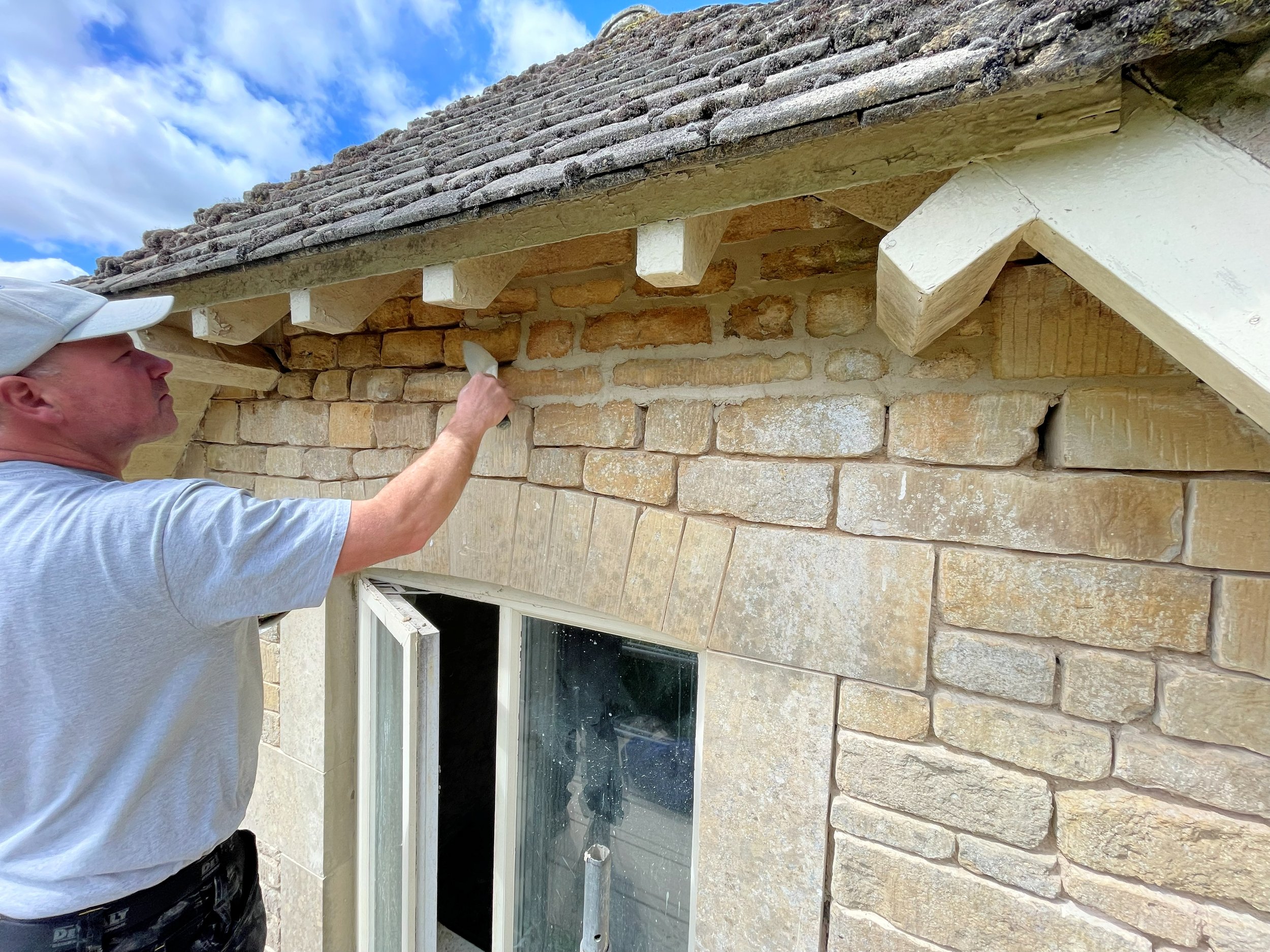 A man working on Cotswold Stone wall under roof, installing or repairing wood trim and lime pointing.