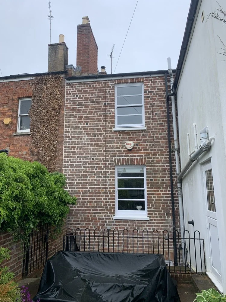 Backyard of brick house with two windows, security alarm, green bush, and covered furniture, next to neighboring white house.