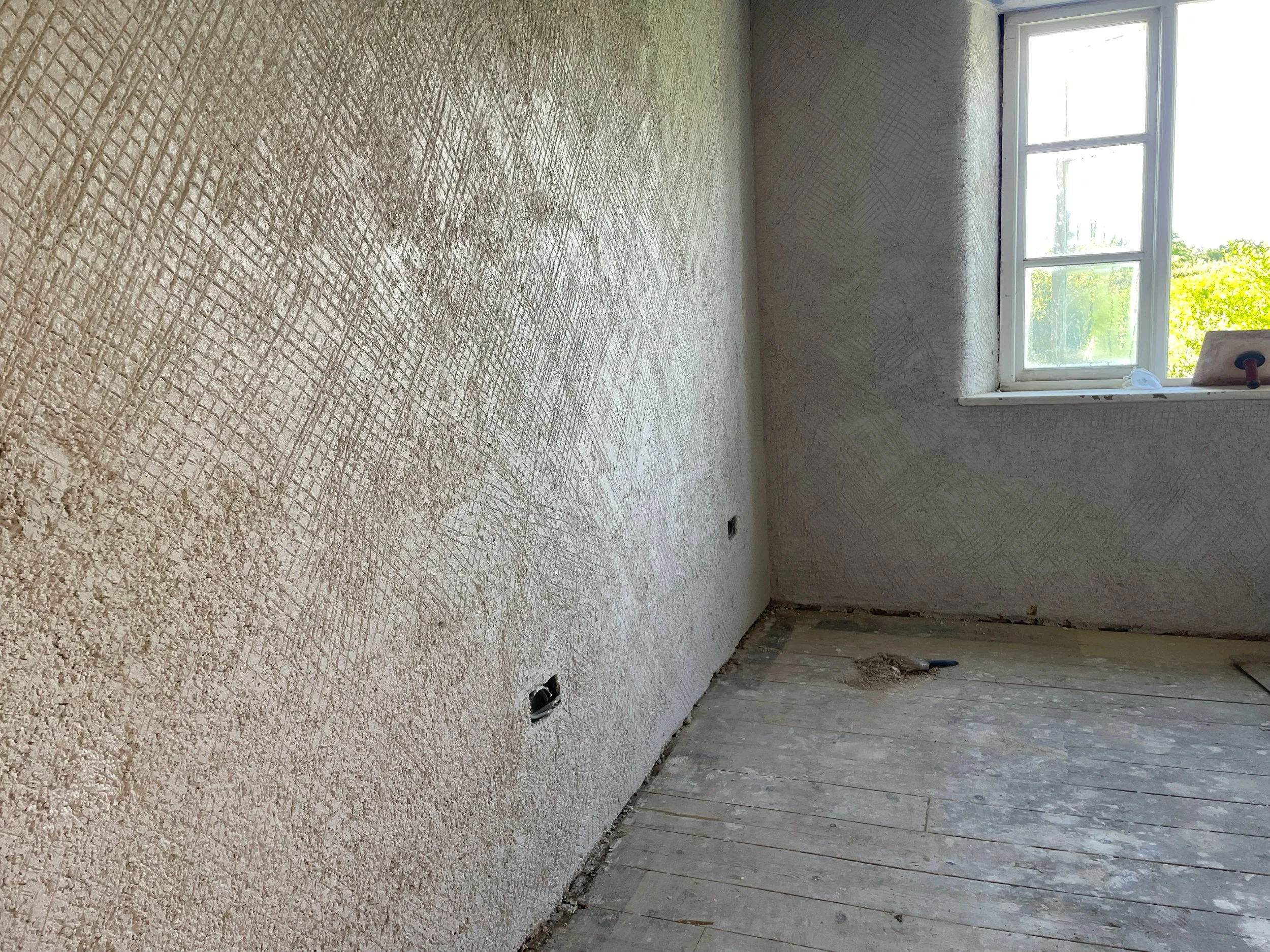 An empty room under renovation with a textured wall, a window with a ledge, and exposed floorboards.
