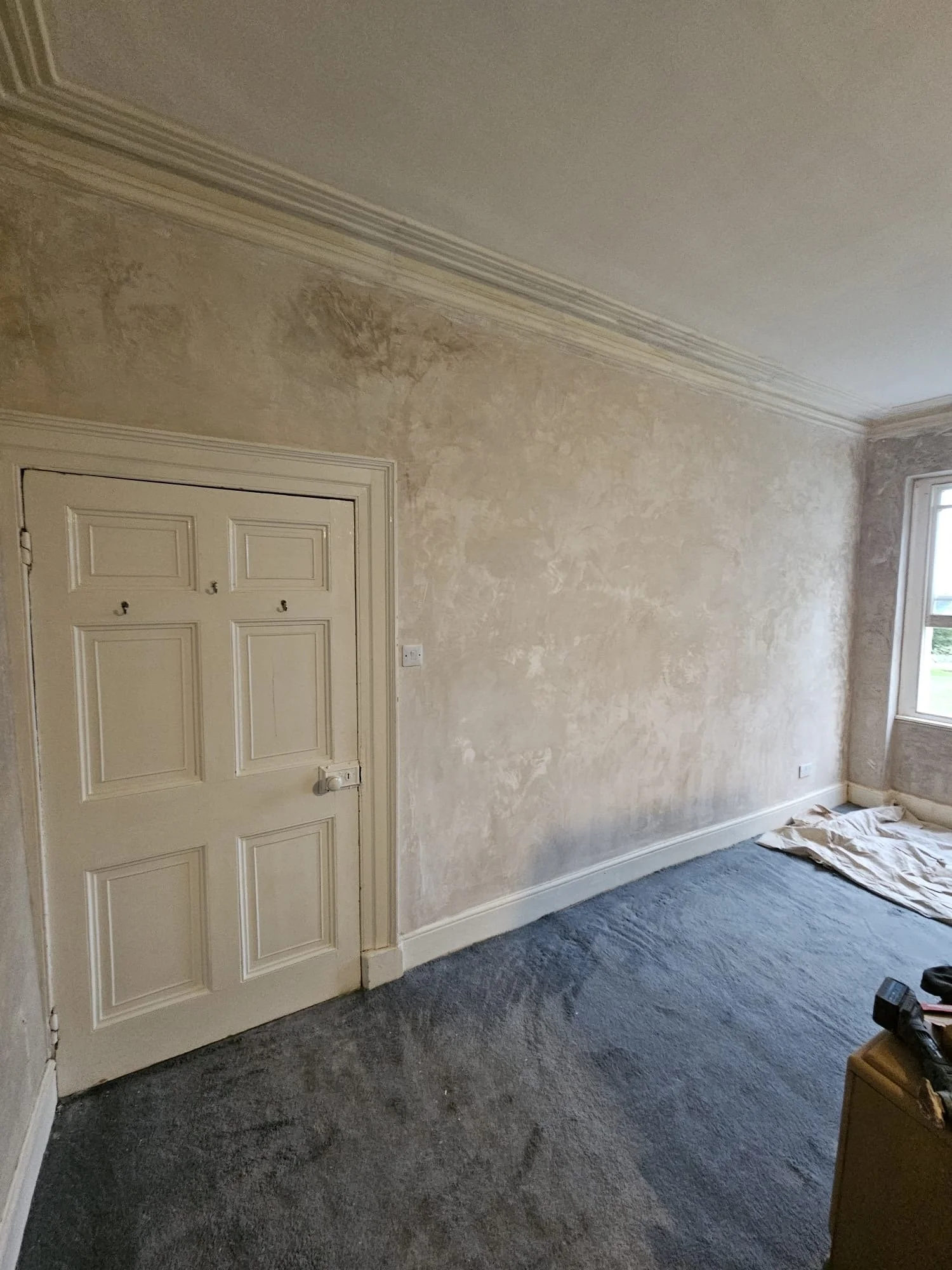 Room with beige textured walls, a closed white door with multiple panels, a window on the right side, and dark gray carpeted flooring. There is a piece of fabric or paper on the floor near the window and a small piece of furniture or object on the bo