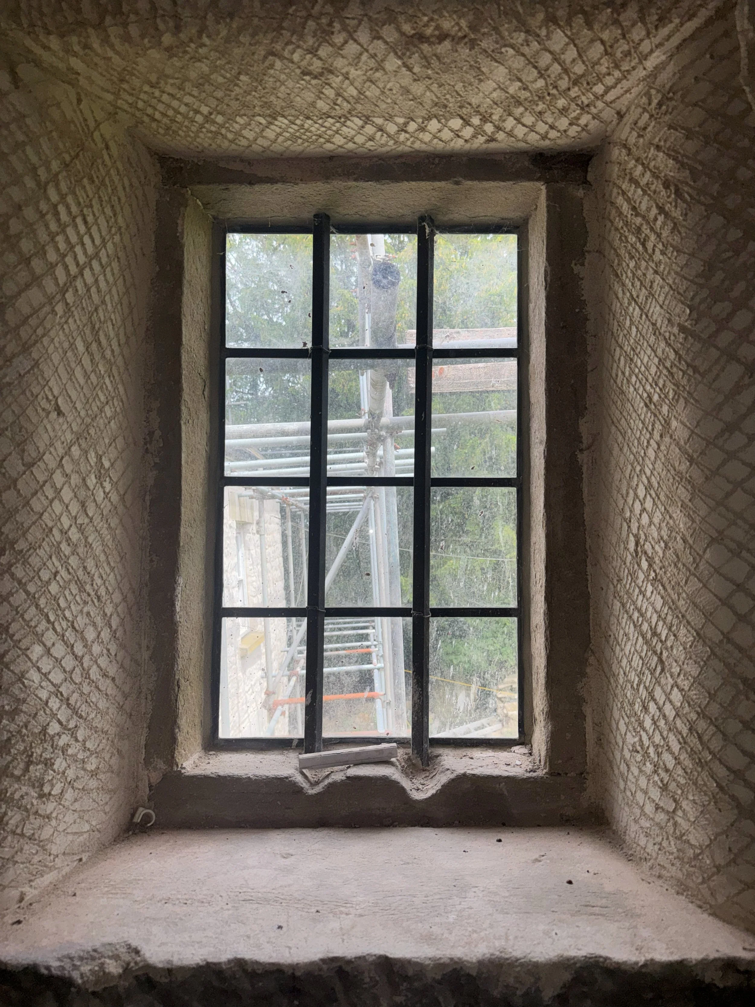 View through a small, rectangular window under construction or renovation, with new brick walls and scaffolding outside, showing trees and sky in the background.