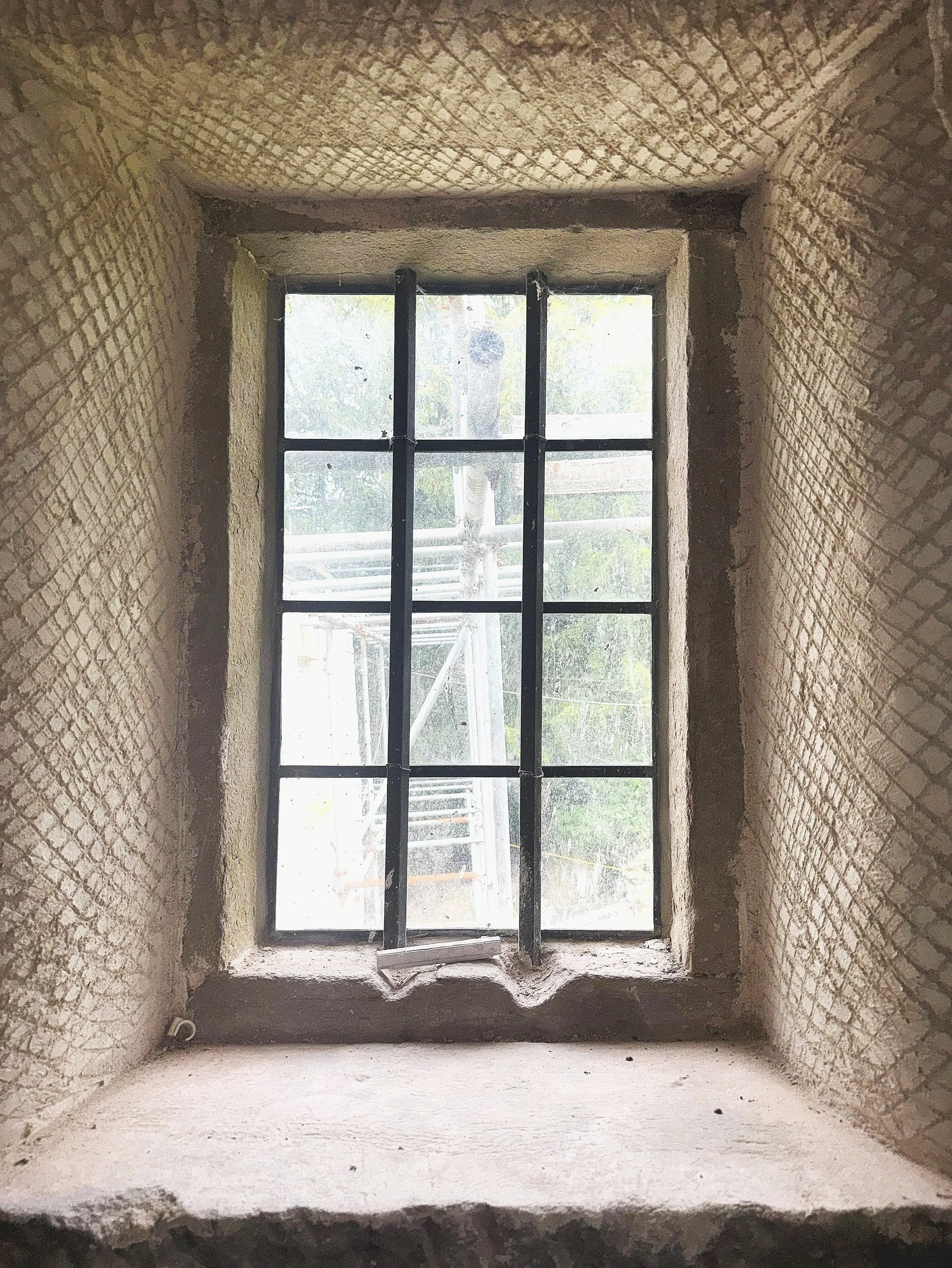 View from inside a window with black metal bars, looking out at a tree and some construction or scaffolding outside. The window is set in a thick brick wall with a rough, unfinished interior.