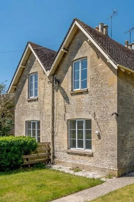 Photo of a listed two-story Cotswold stone house with a steep roof, four windows, and a small front yard with green grass and a wooden fence.