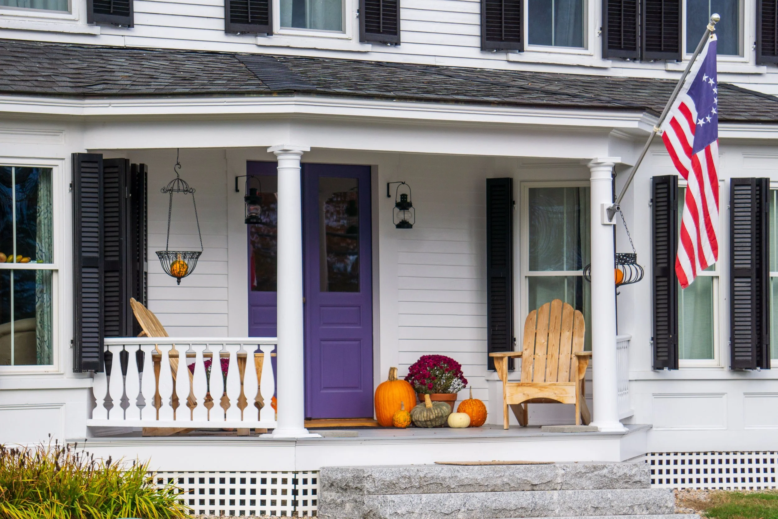 Front porch of a large white home, with a flag out front and several pumpkins on display.