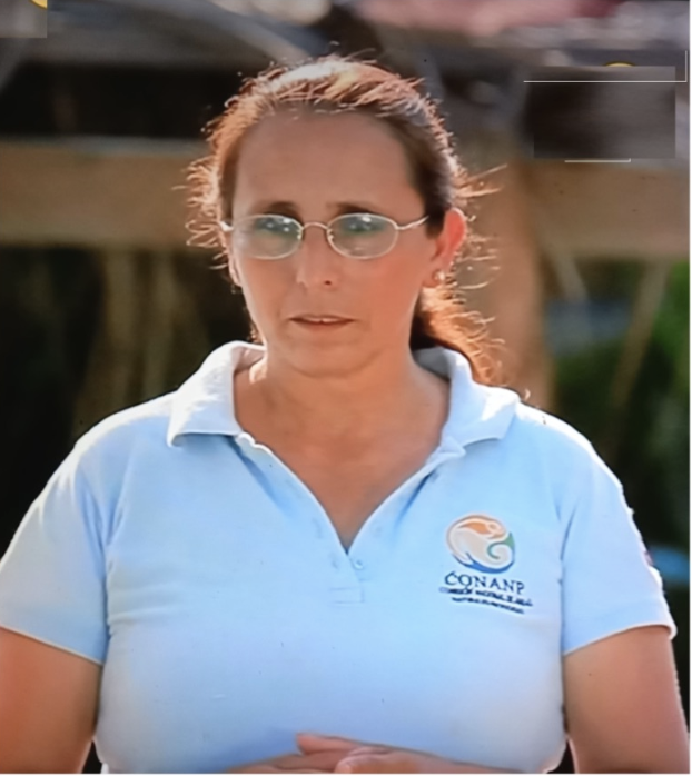 A woman with brown hair, wearing glasses and a white polo shirt with a logo that says 'CONANP.' She appears outdoors, with a blurred background of wooden structures.
