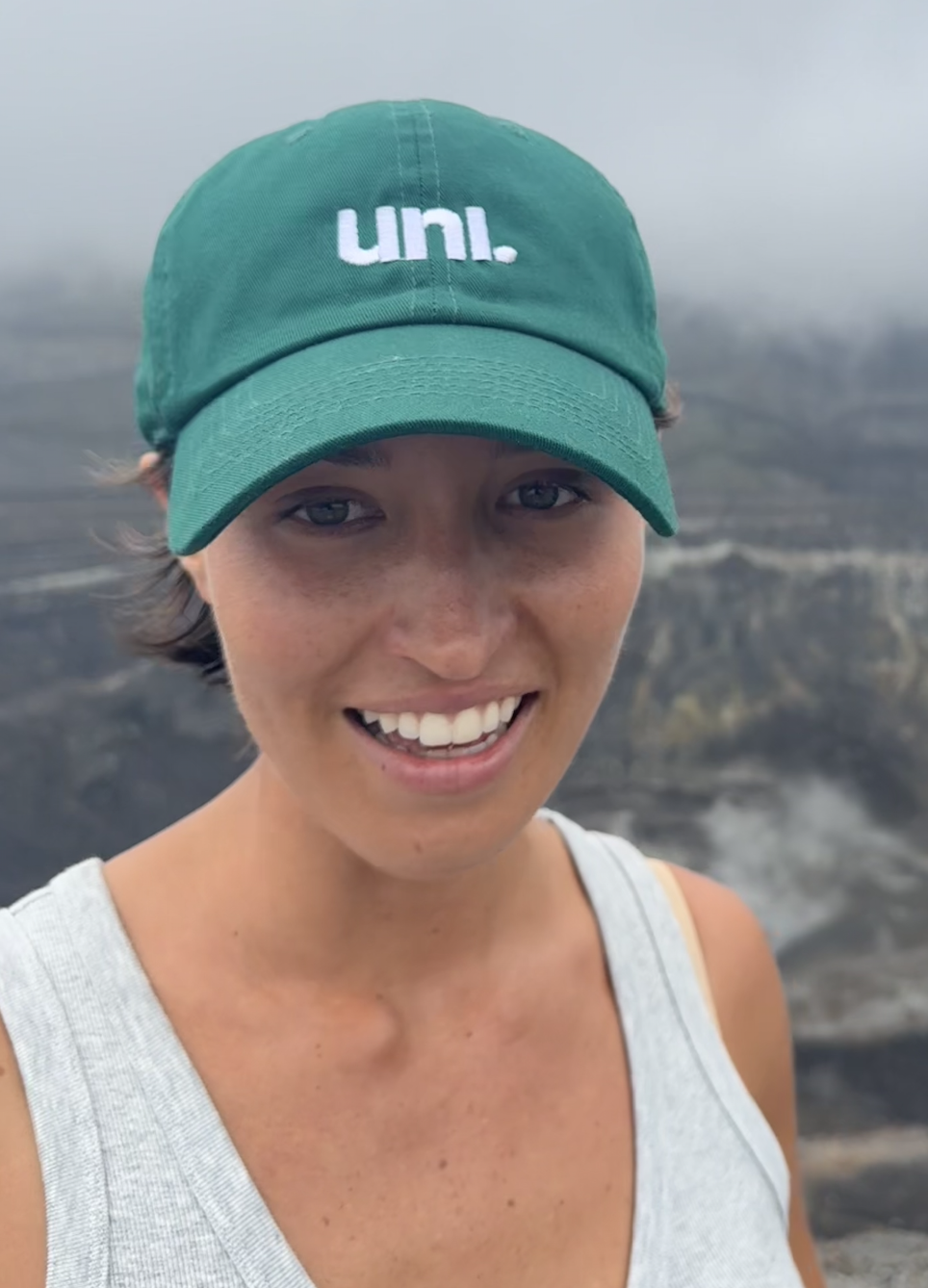 A woman smiling outdoors wearing a green cap with white text, with a blurred background of what appears to be a waterfall or rocky landscape.