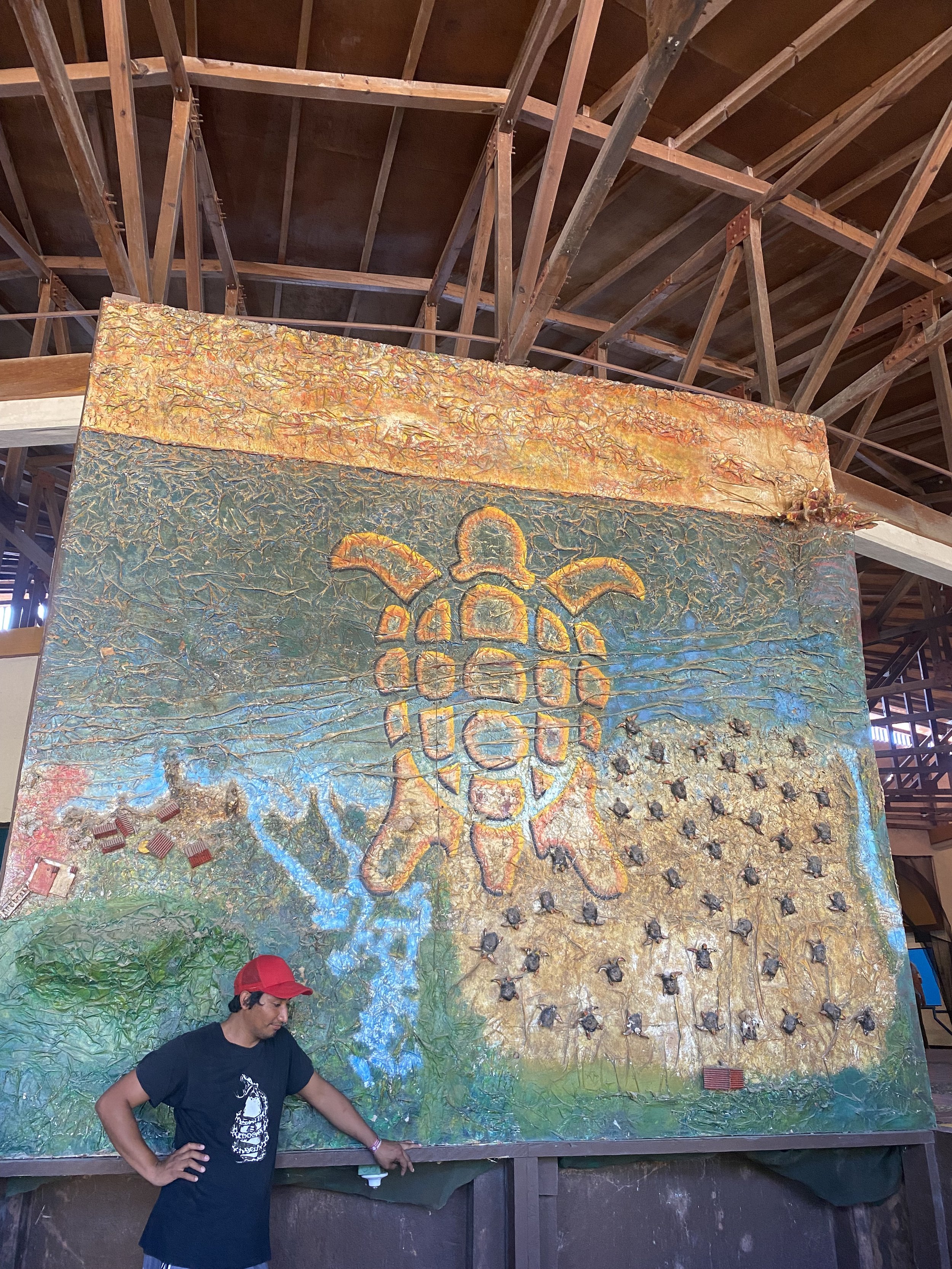 A man wearing a black t-shirt and red cap standing in front of a large outdoor art installation. The artwork features a textured surface with various colors and a prominent depiction of the number 300, surrounded by stars and a bee. The wooden beams overhead indicate an outdoor or semi-outdoor setting.