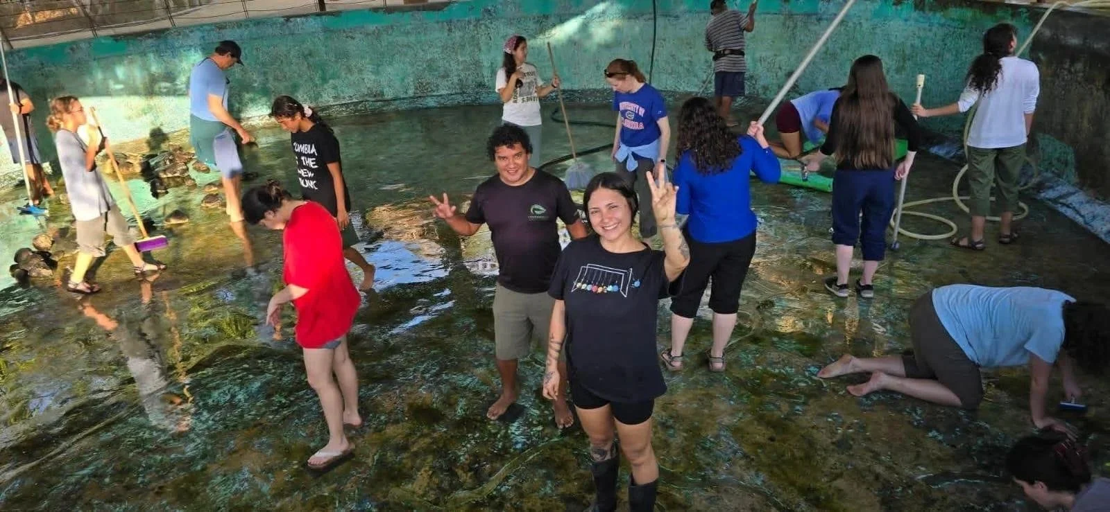 People exploring an indoor shallow water tank, some collecting rocks or using tools, and two women in the foreground smiling and making peace signs.