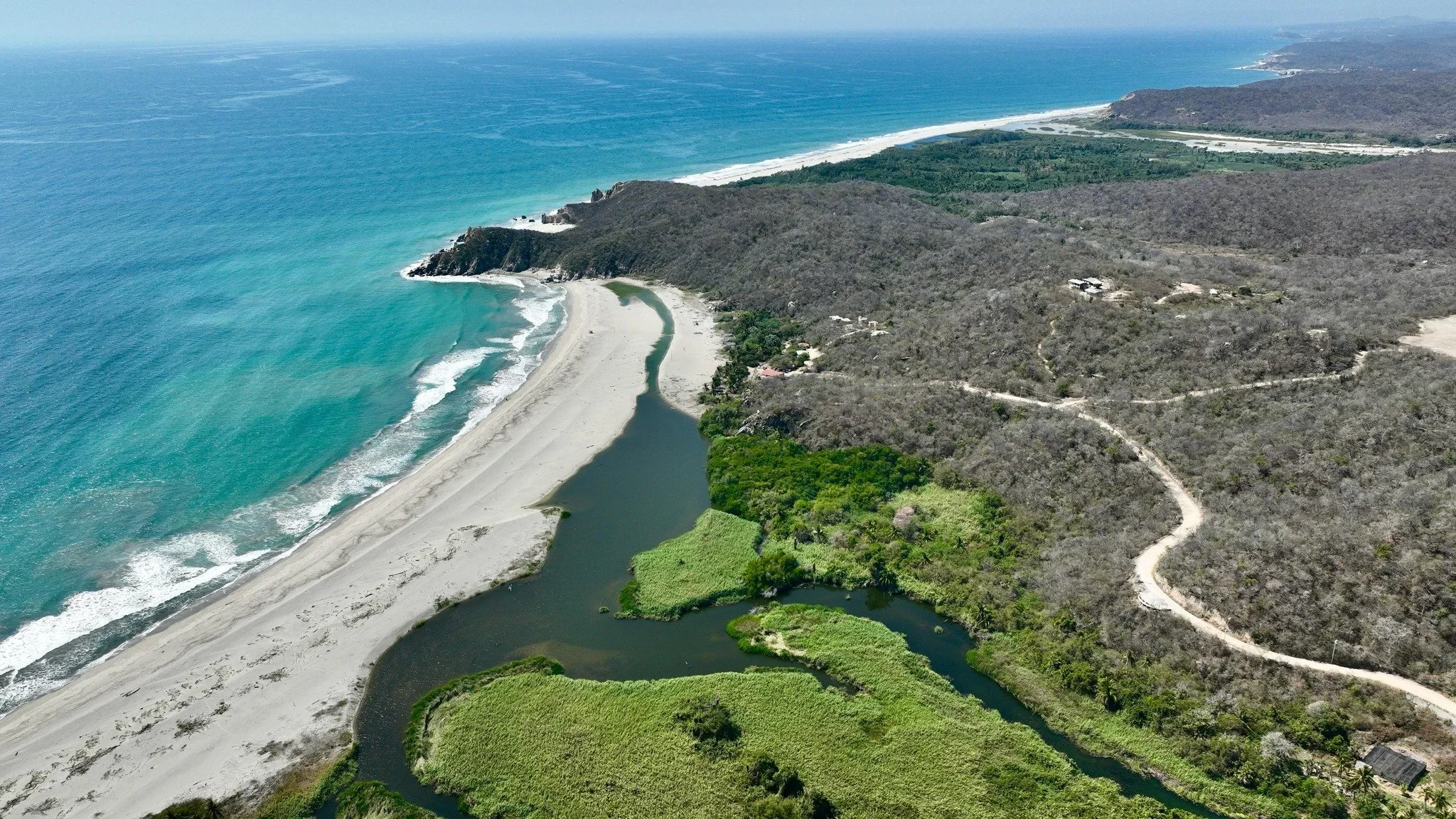 Aerial view of a scenic coastline with a beach, ocean, and hilly terrain, featuring a river or creek winding through green vegetation.