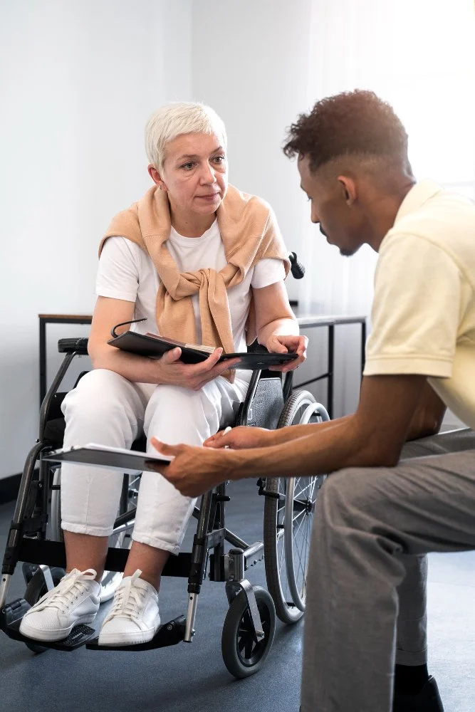 A woman in a wheelchair and a man are having a serious conversation in a bright room, possibly a medical or counseling setting.