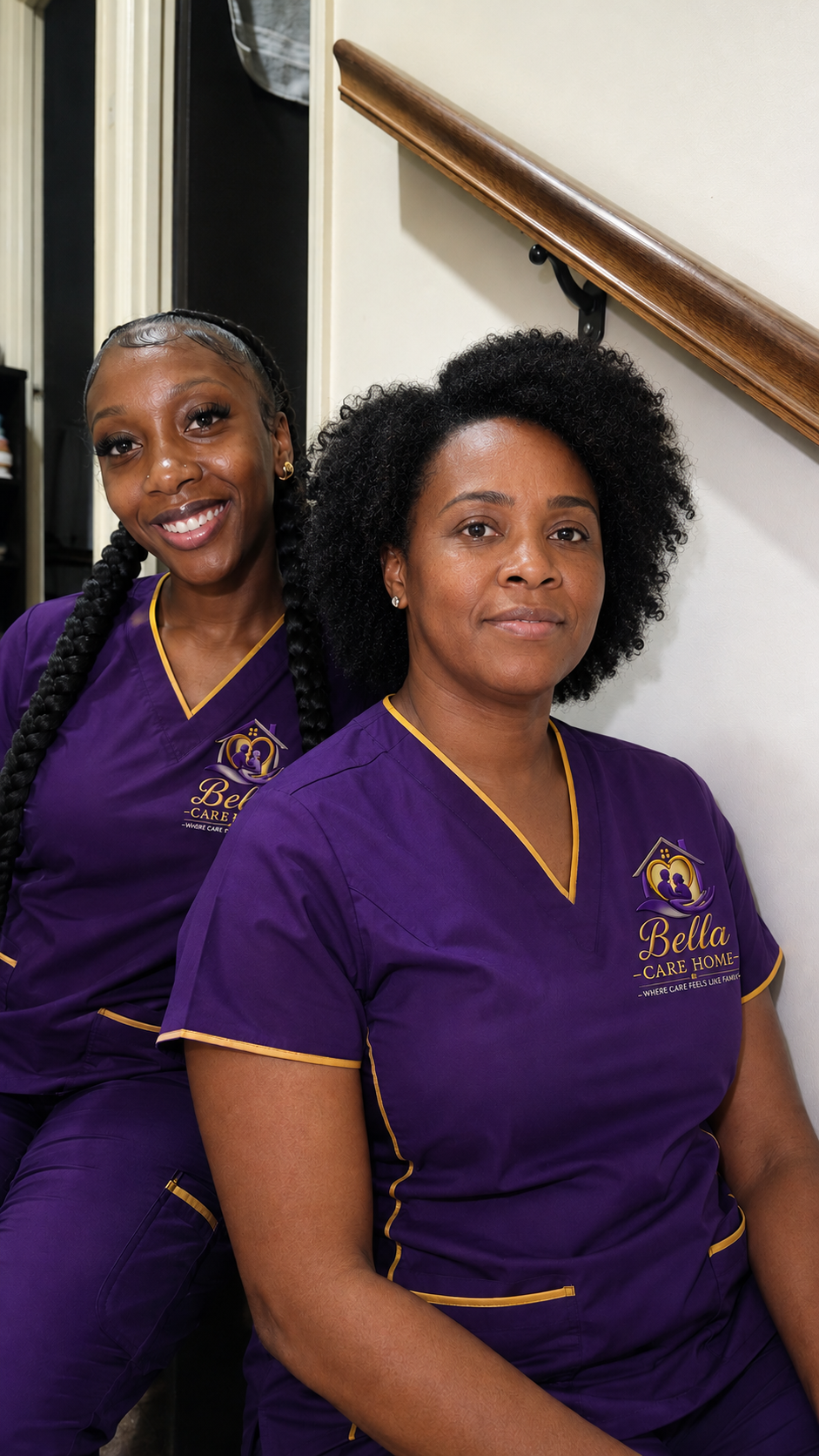 Two women in purple uniforms with yellow trim, both with natural hairstyles, standing near a staircase and smiling at the camera. Their uniforms display the logo and name 'Bella Care Home'.