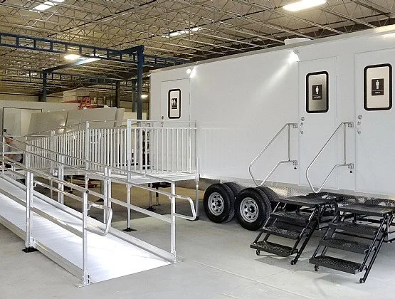 Accessible portable restroom trailer with wheelchair ramp and stairs in an industrial warehouse setting.