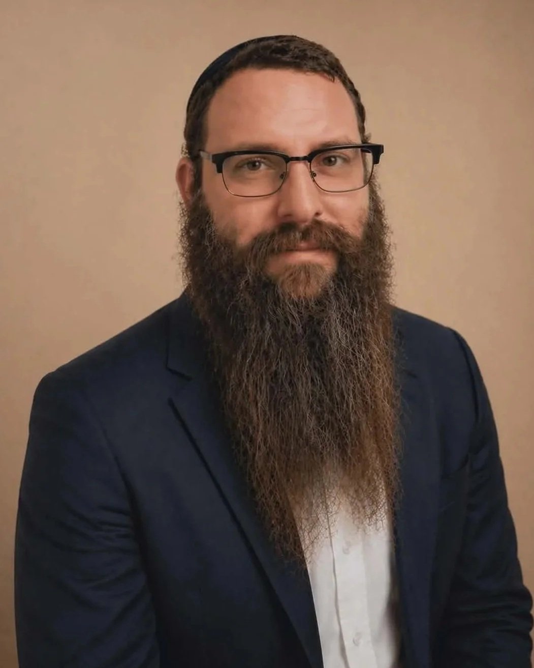 A man with glasses, a long beard, and short hair wearing a navy blazer and white shirt, posing in front of a beige background.