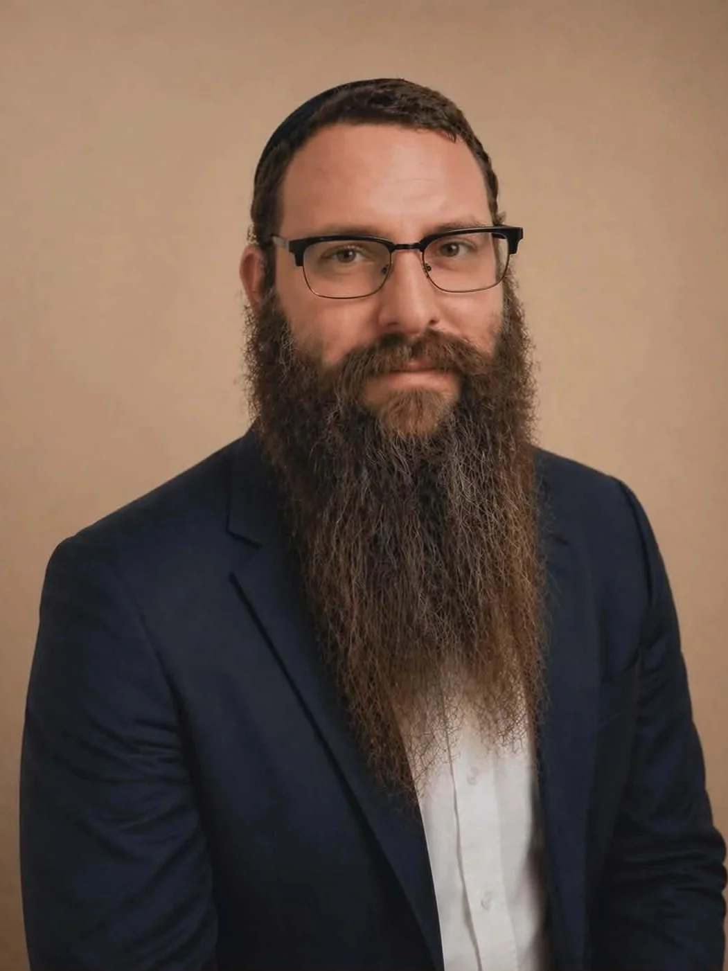 A man with glasses, a beard, and mustache, wearing a black blazer over a white shirt, posing against a beige background.