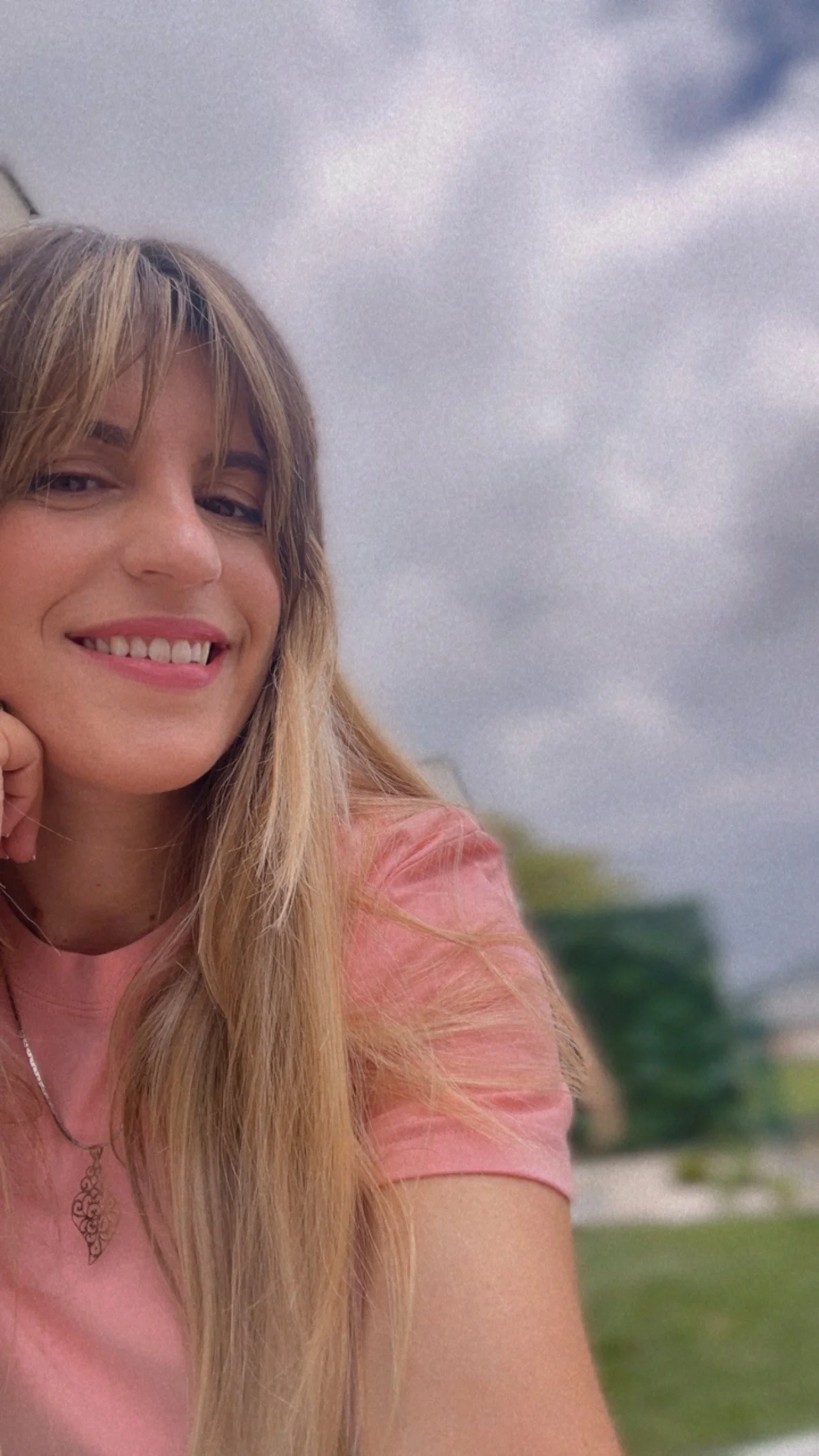 Mulher de cabelo louro longo, sorrindo, usando uma camiseta rosa, ao ar livre, com céu nublado ao fundo.