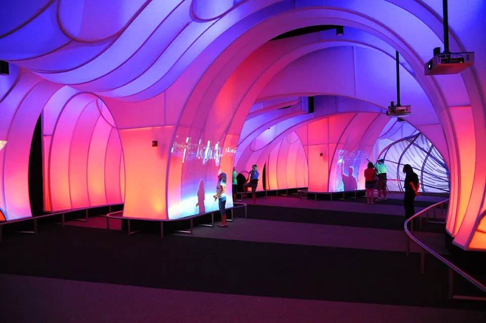 Colorful, futuristic corridor with curved walls illuminated in pink, purple, and blue lights, with several people engaging with interactive displays.