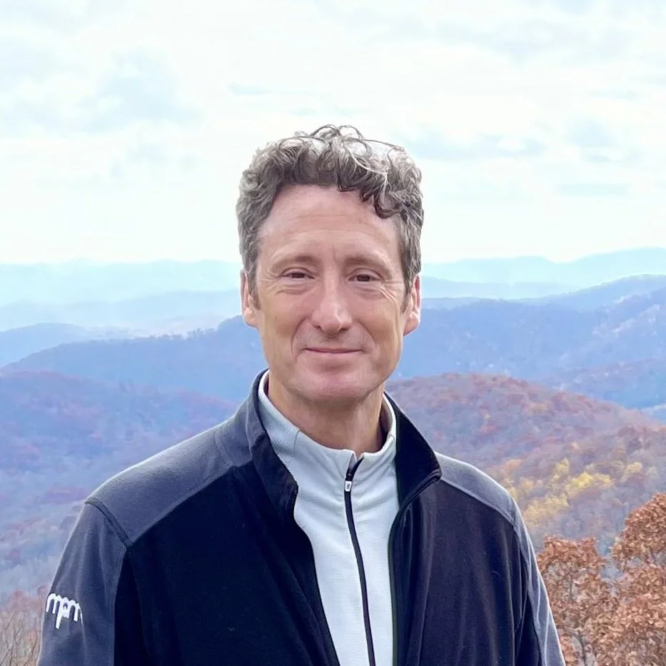 Man standing outdoors with a backdrop of mountains and trees in autumn colors.