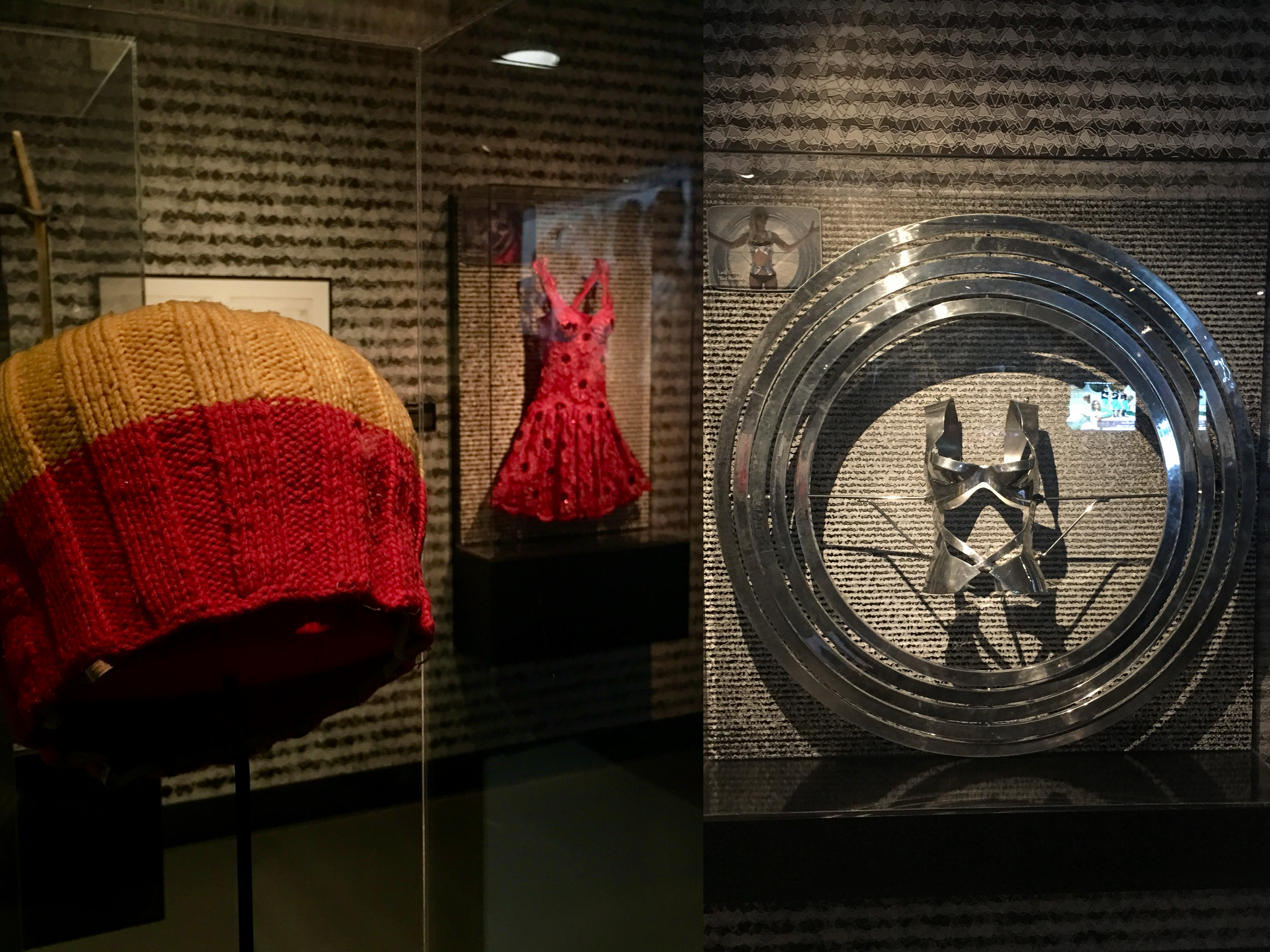 Museum display with a knitted object in shades of orange, red, and yellow, a framed figurative artwork in the background, a small image of a woman in a sports bra and shorts, and a large metallic star sculpture with concentric rings behind it.