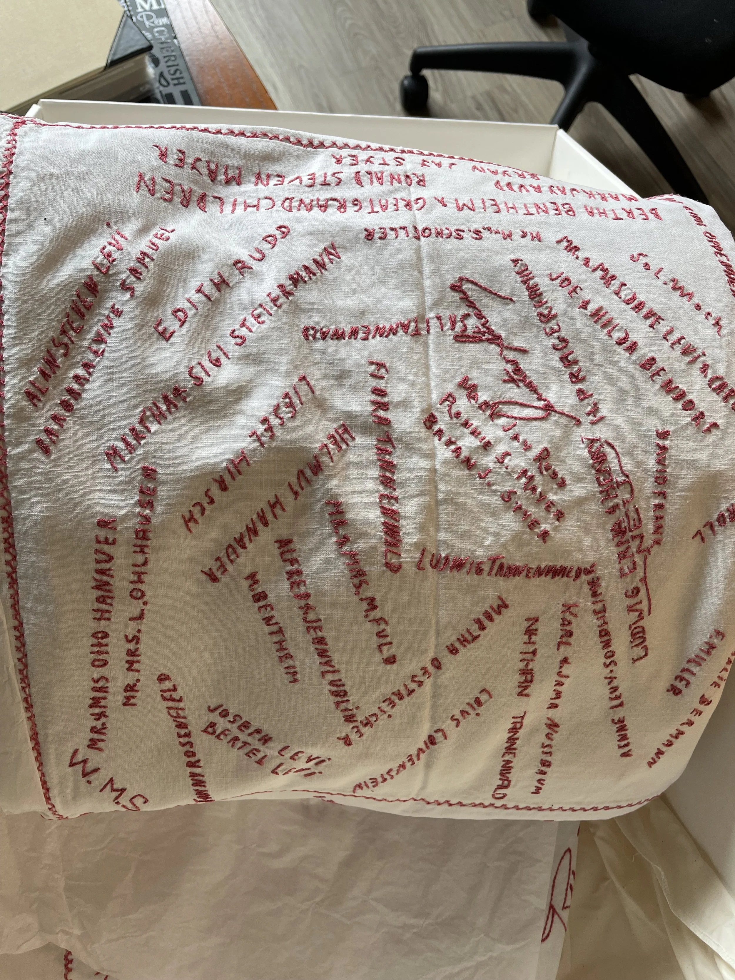 A white cloth napkin with red embroidered text arranged in a spiral pattern. The embroidery includes the words "SALUTE" and various other text, along with decorative lines and symbols.