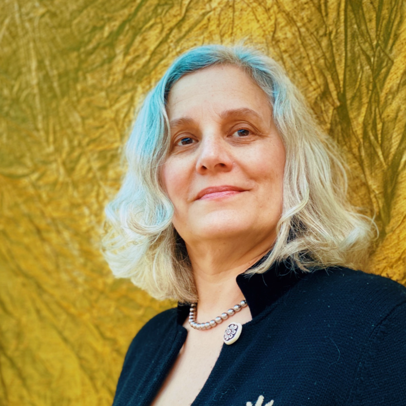A woman with shoulder-length gray hair with blue streaks, smiling, wearing a black top and a pearl necklace, standing in front of a yellow textured background.