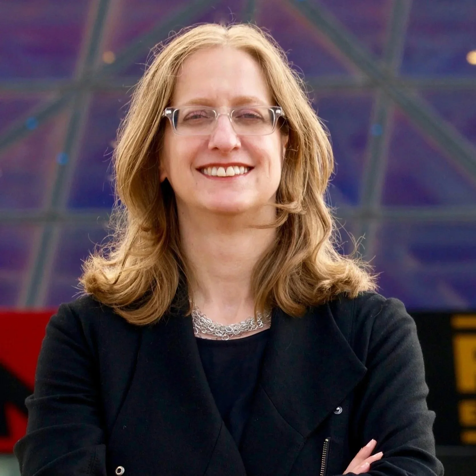 A woman with light brown, shoulder-length hair smiling in front of a colorful, geometric patterned background. She is wearing glasses, a black top, and a silver chain necklace.