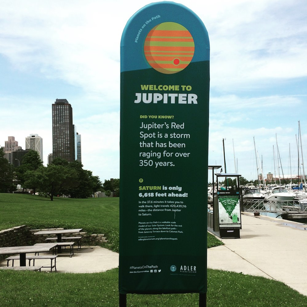 An informational sign at a park by the waterfront featuring details about Jupiter and Saturn, with a cloudy sky and city skyscrapers in the background.