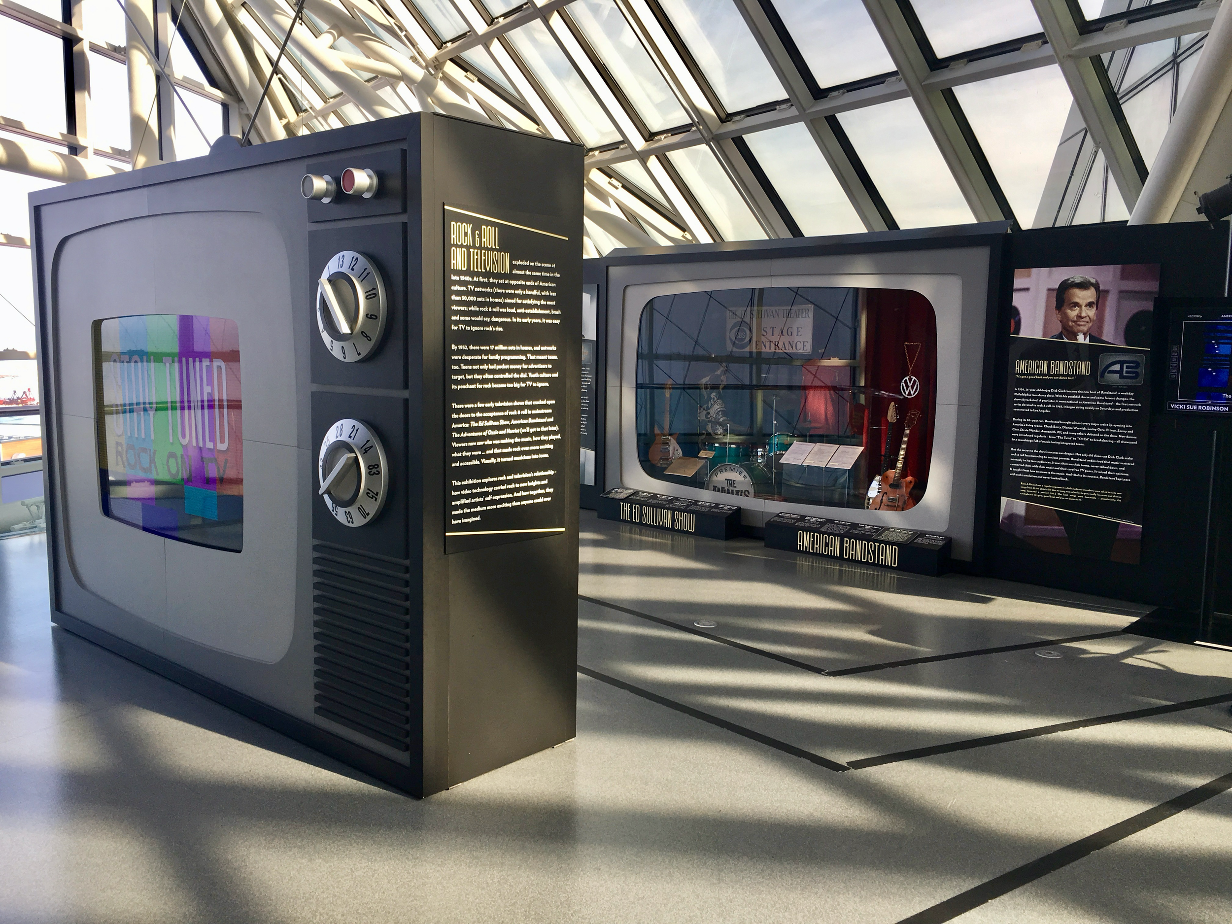 An indoor exhibit space with a large display case and informational panels. The case contains musical instruments and memorabilia, including guitars and a necklace. The lighting is natural, coming through a glass ceiling and windows.