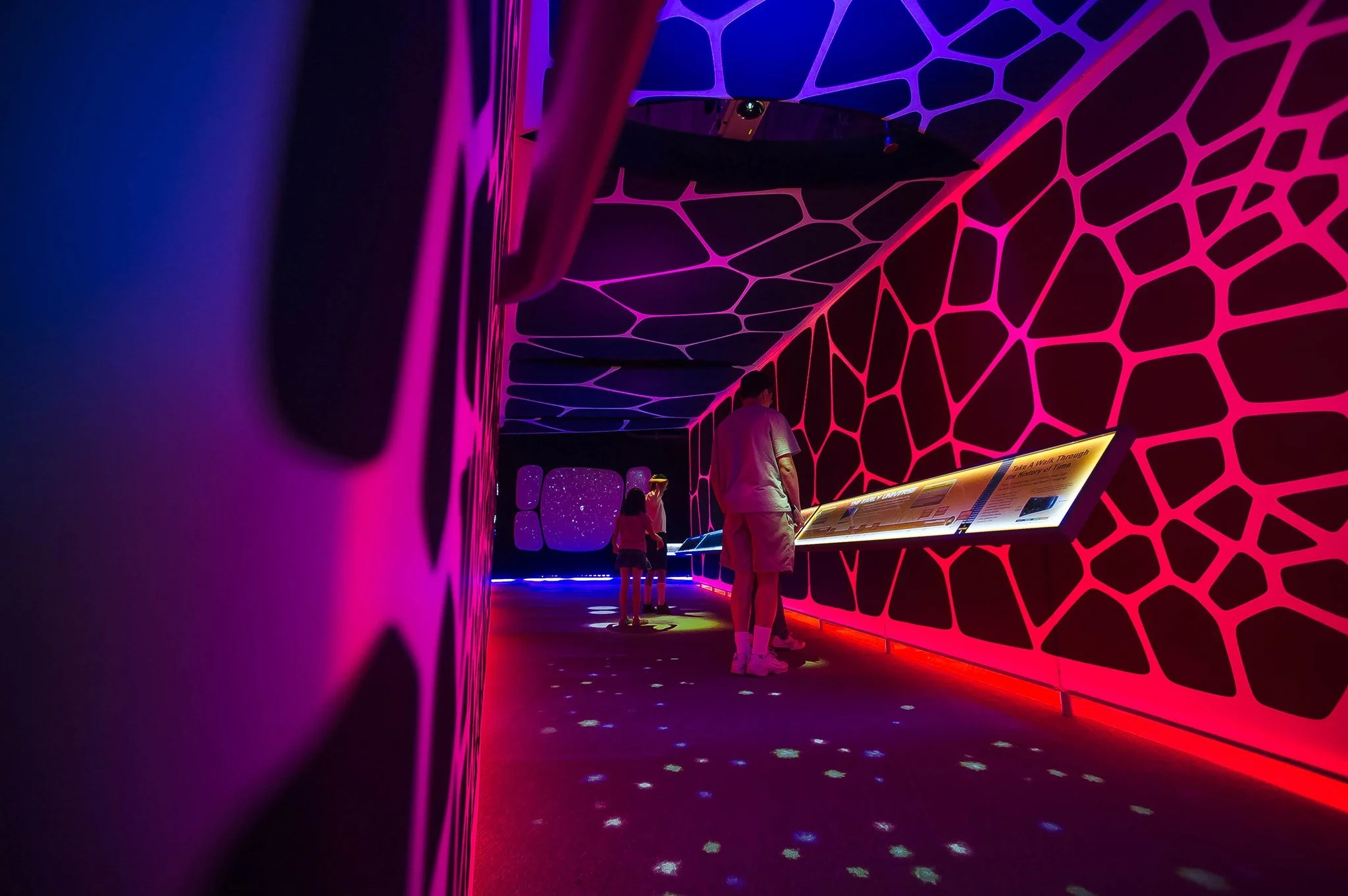 People exploring a dark room illuminated with vibrant neon lights and geometric patterns, with informational display panels.