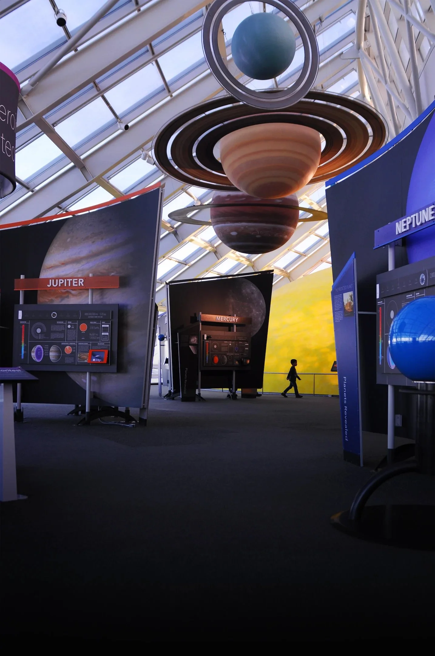 Interior view of an exhibition space with a large planetary model hanging from the ceiling, showing Saturn with rings, and posters of other planets like Jupiter and Neptune on display.