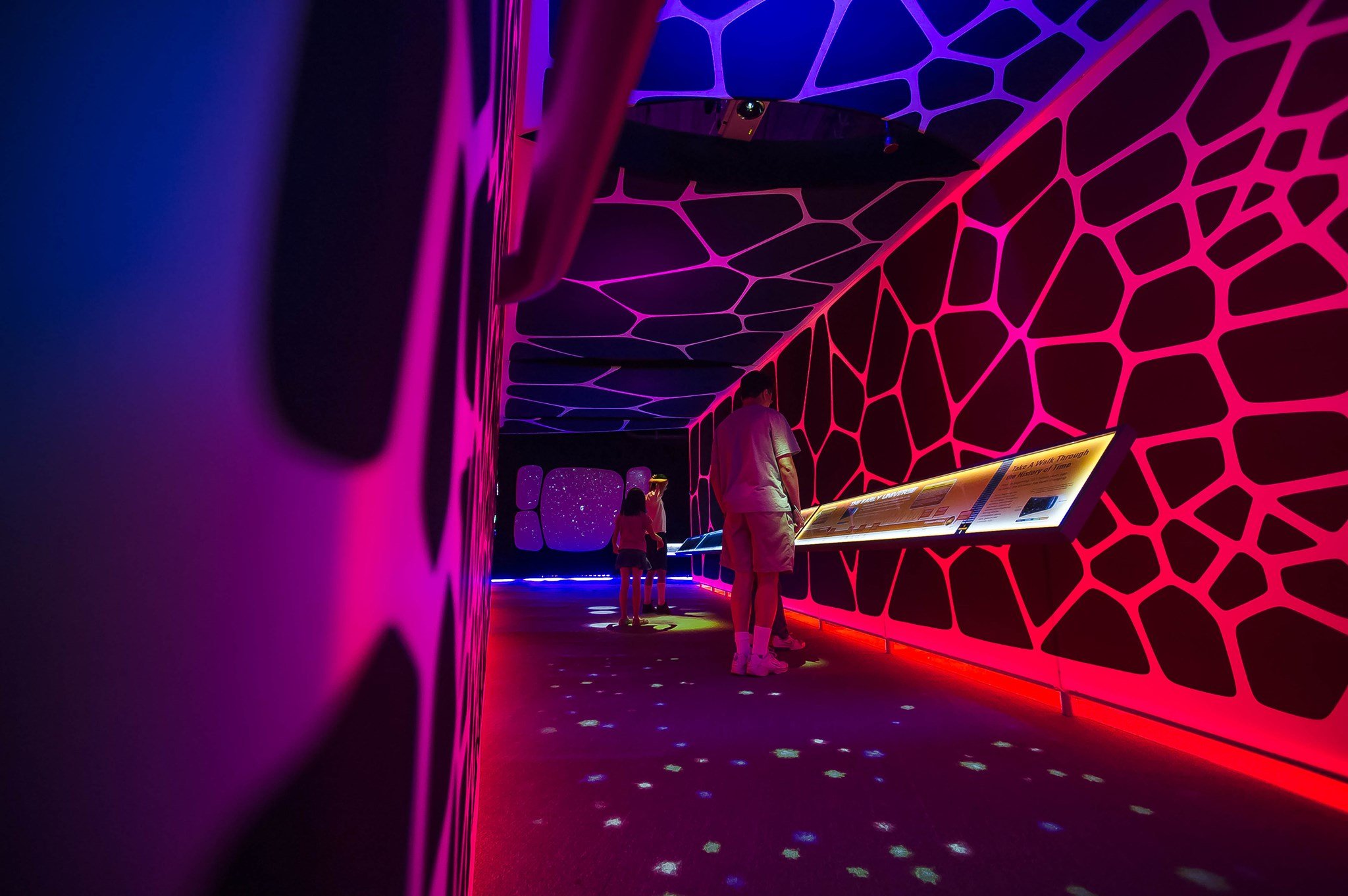 People exploring a vibrant, neon-lit exhibit with glowing geometric patterns and a touch screen display.