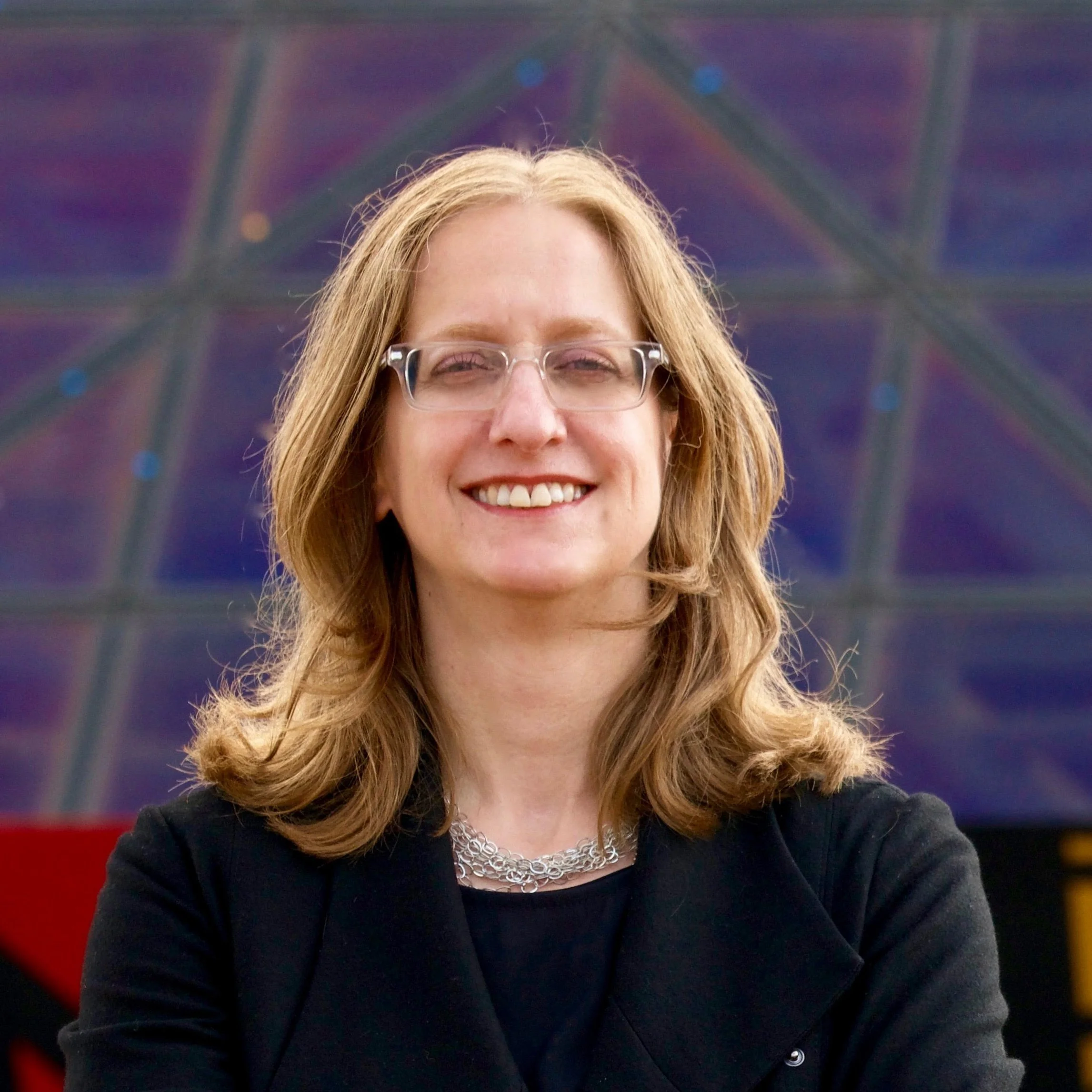 A smiling woman with blonde hair wearing glasses and a black top, standing in front of a blurred background with multicolored abstract patterns.