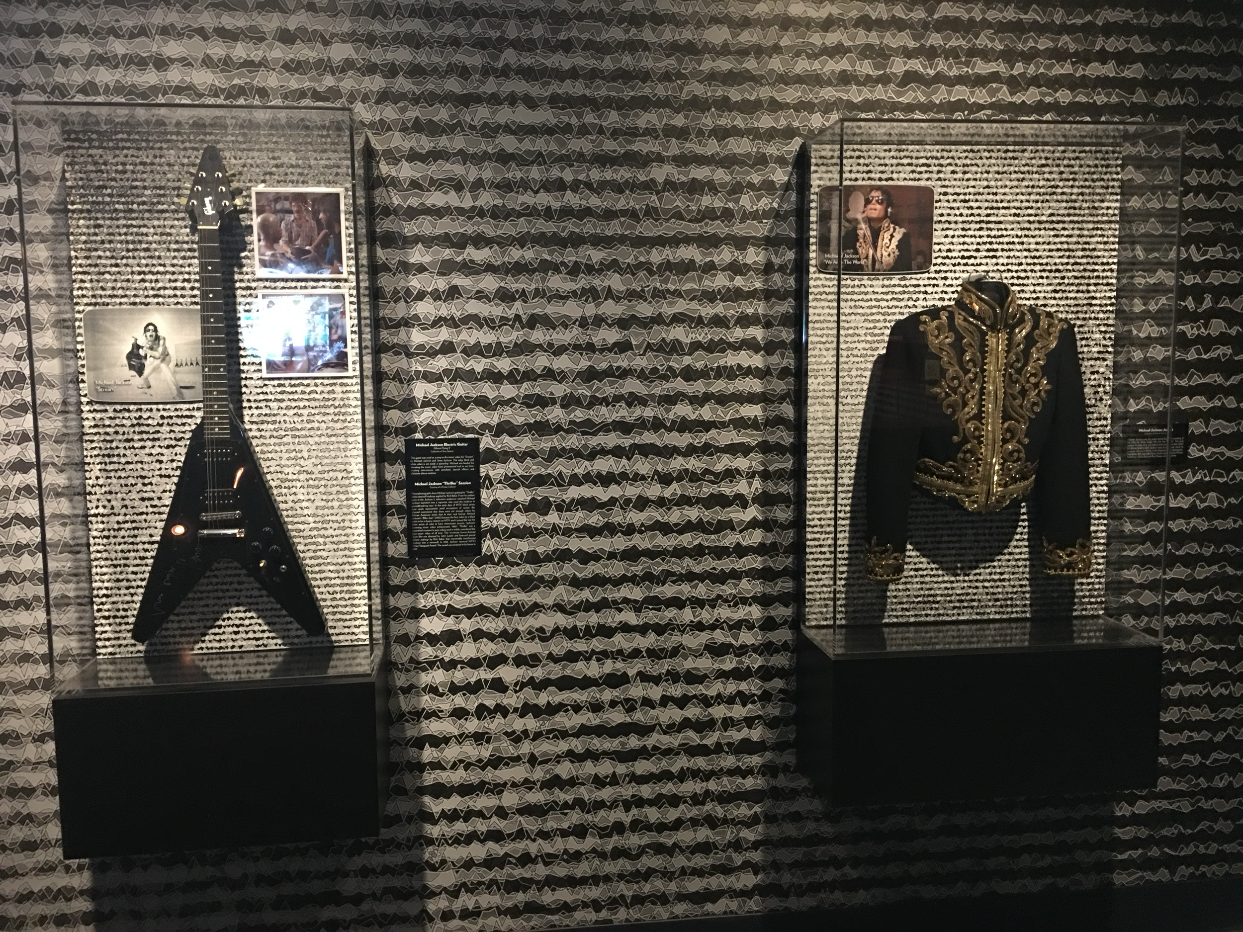 Display case featuring a black electric guitar, framed picture of Michael Jackson, and a black jacket with gold embroidery on a patterned backdrop.
