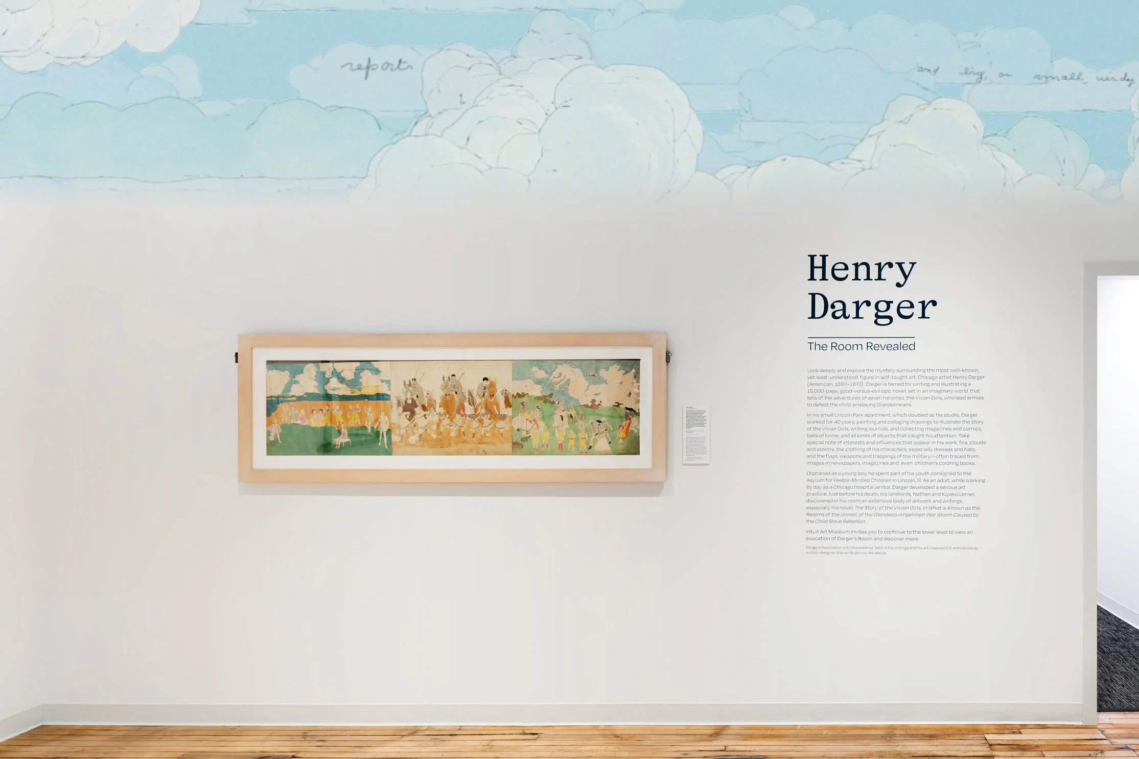 An art exhibit featuring a large framed painting depicting multiple figures and scenes in vibrant colors. The artwork is hung on a white wall with accompanying descriptive text, and the room has hardwood floors and white walls with a cloud mural near the ceiling.