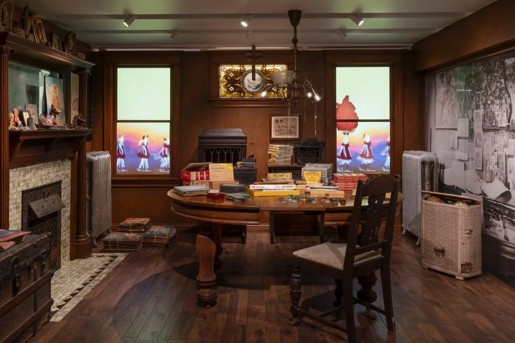 A cluttered office or study room with wooden walls and furniture, two windows with display screens, a fireplace, and stacks of books, papers, and magazines on the table and shelves.