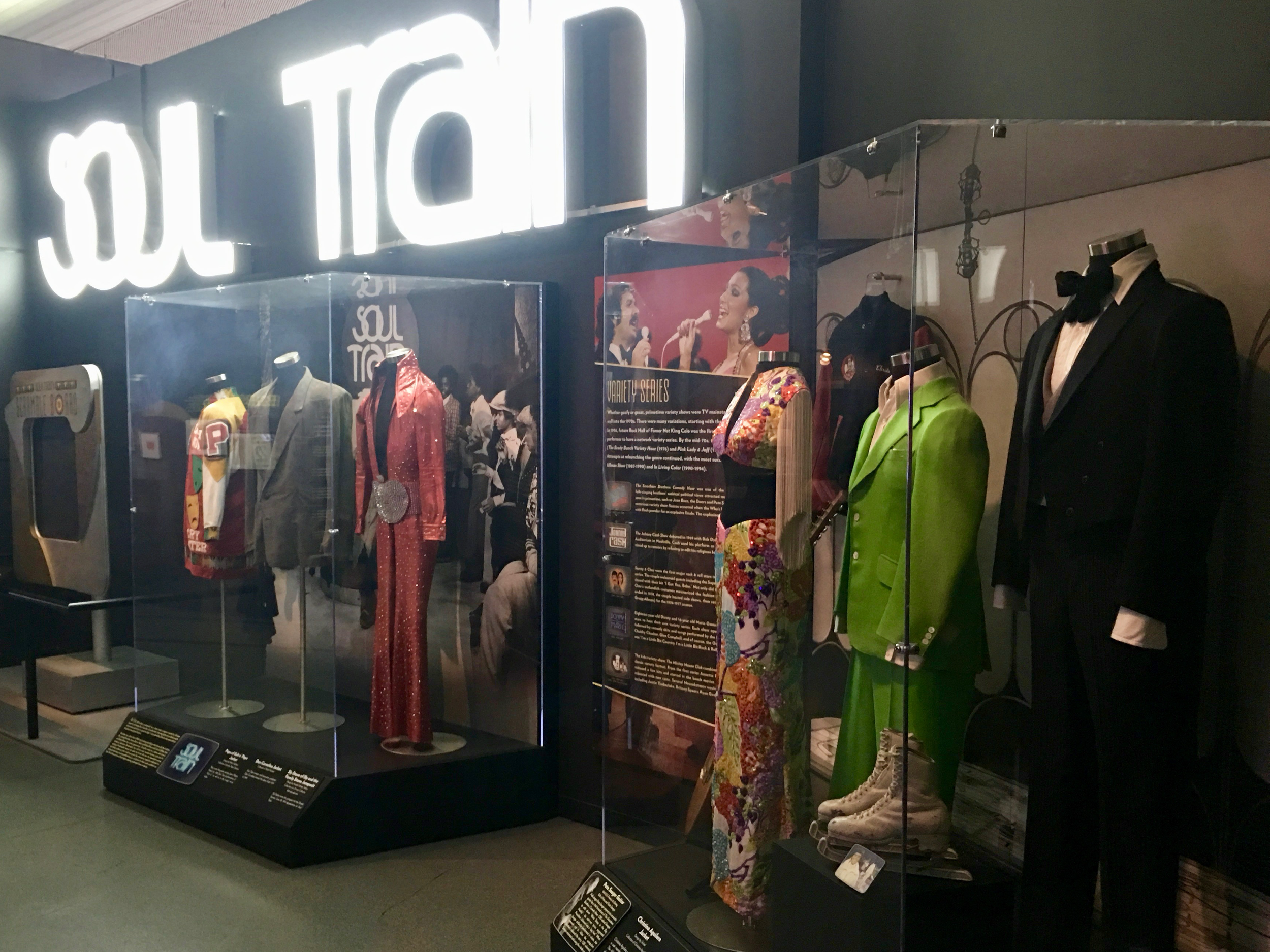 Exhibit of vintage costumes including a gray suit, a red dress, a colorful floral dress, a bright green suit, and formal black tuxedo with bow tie, displayed on mannequins behind glass at a museum.