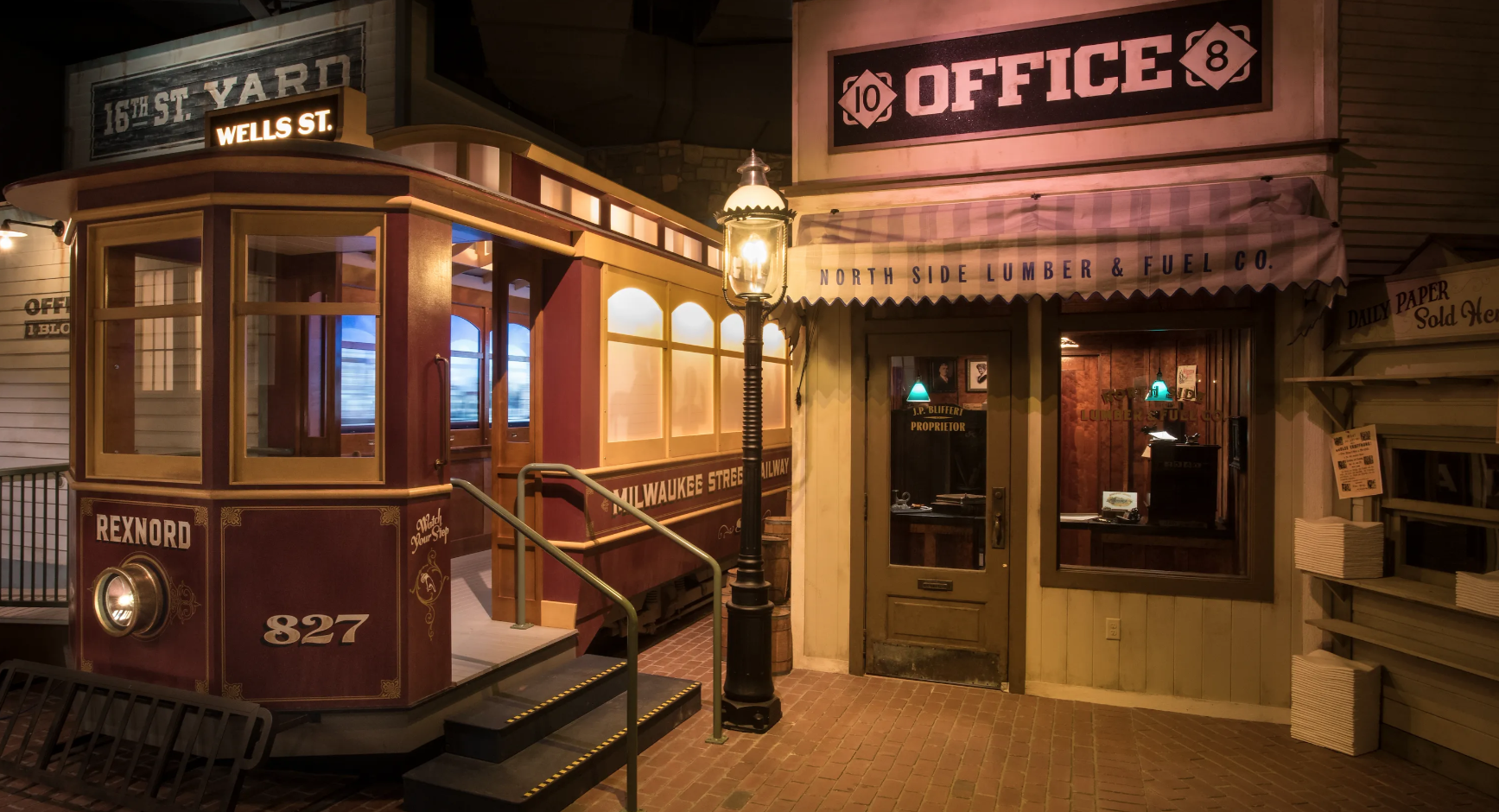 Vintage street scene with a red trolley car, a lamp post, and various storefront signs including '16th St. Yard,' 'Wells St,' 'Rexnord 827,' 'Milwaukee Street Railway,' and 'North Side Lumber & Fuel Co.' at night.