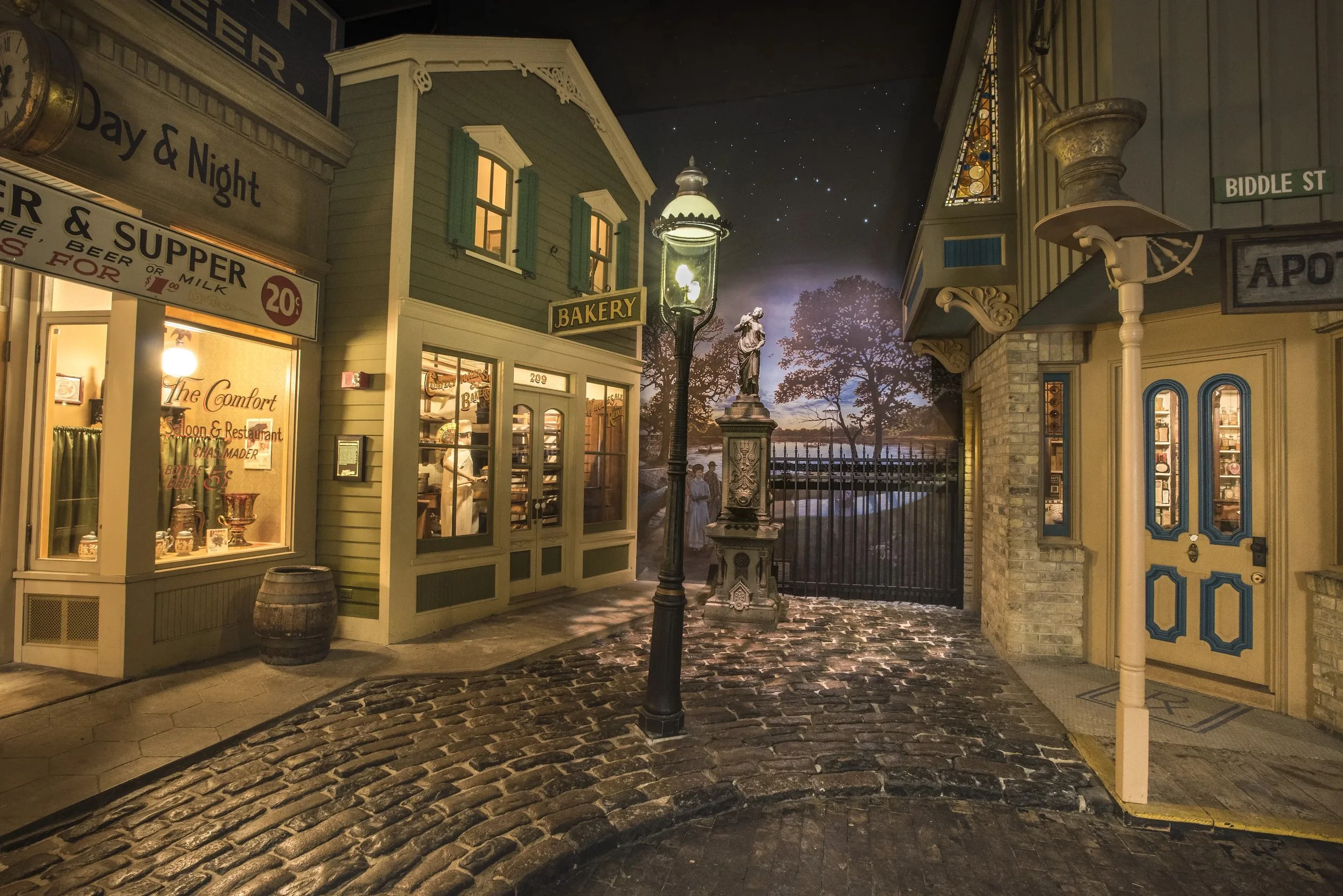 A detailed street scene at night, featuring a vintage-style street lamp, storefronts with signs for bakery and other shops, cobblestone pavement, decorative buildings, and a starry sky with a faint outline of distant trees and water.